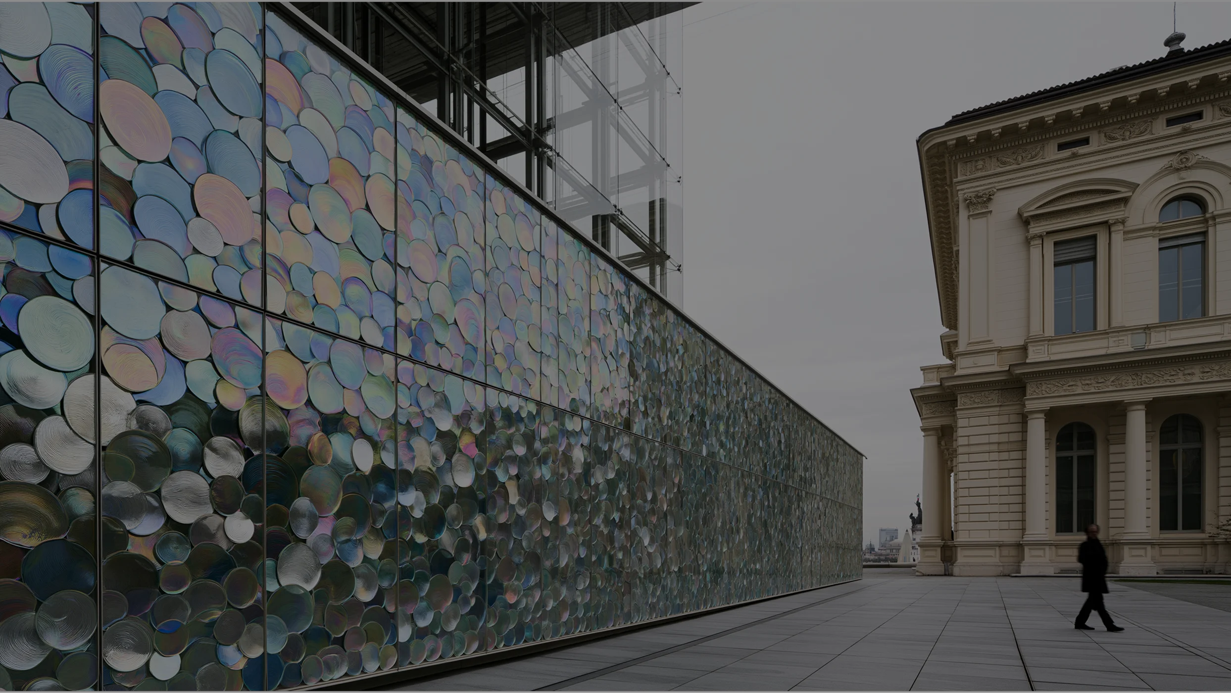 Panoramic view of a modern glass wall covered with iridescent circular elements, next to a classical historical building with a person walking in the forecourt.