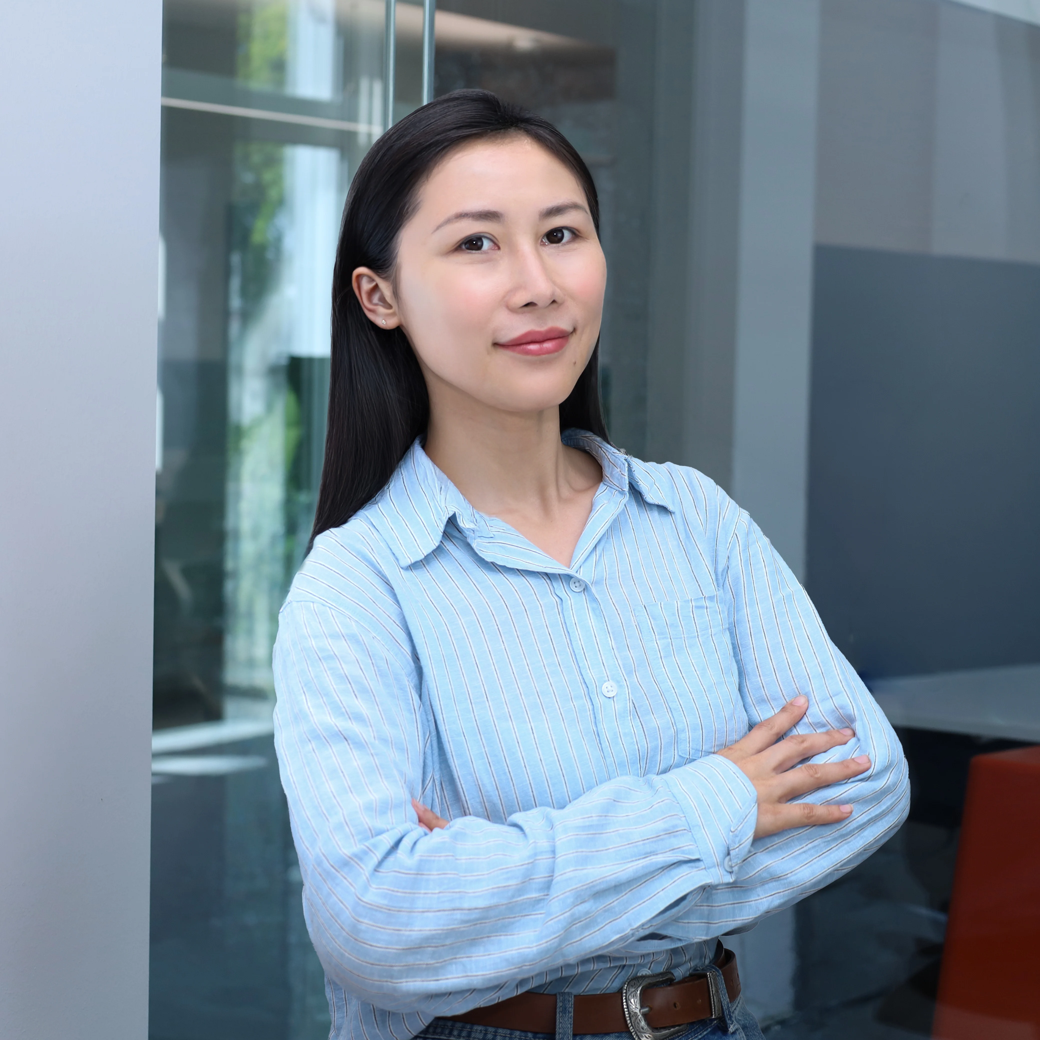 Professional half-length portrait of an Asian woman, Xu He who works at redify as a Senior AI Product Manager with long black hair and a blue striped shirt, posing with arms crossed against a light grey background.