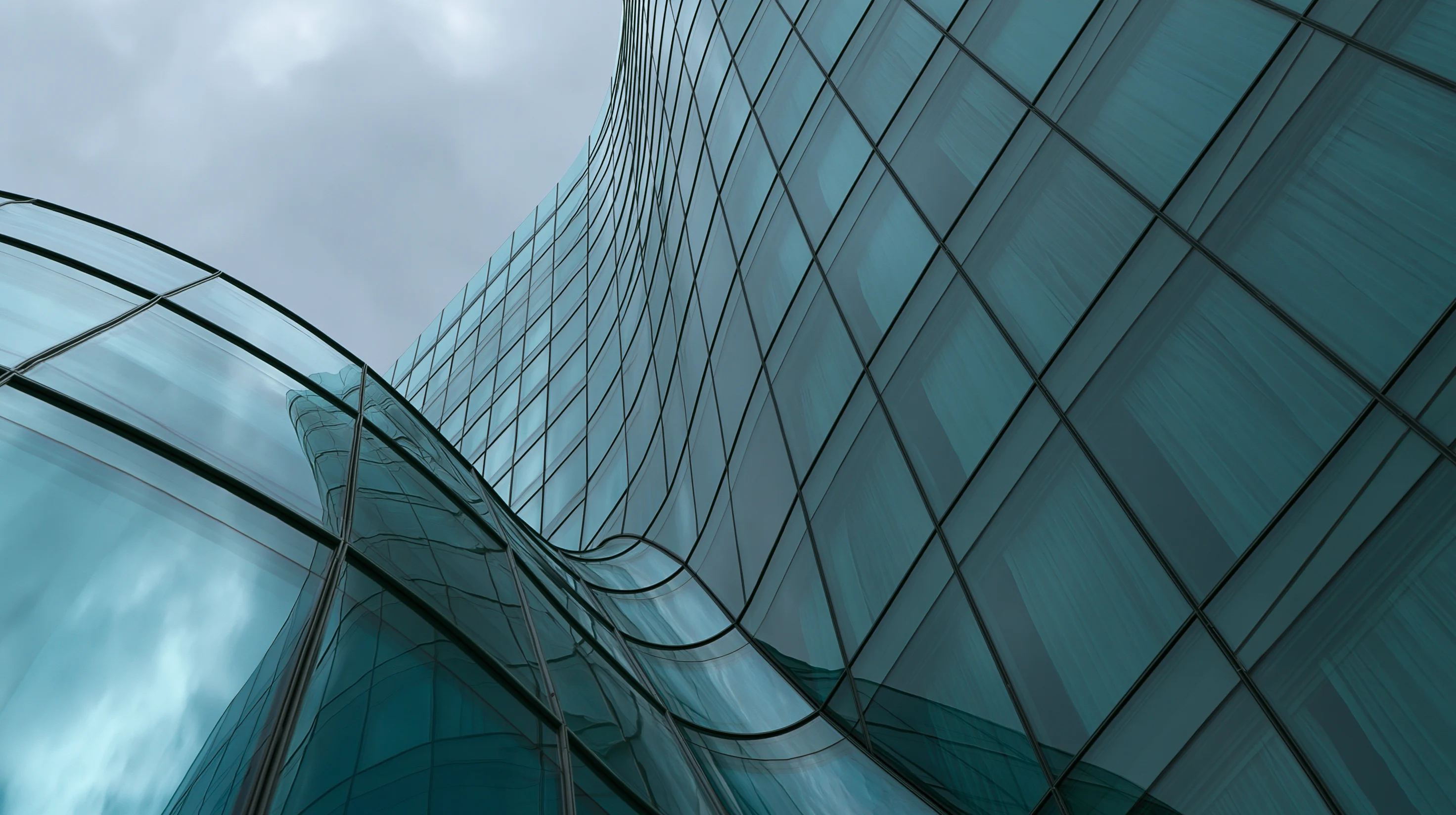 Low-angle shot of a modern skyscraper with a blue-green glass curtain wall. The structure features wavy, sinuous surfaces and reflects an overcast, cloudy sky.