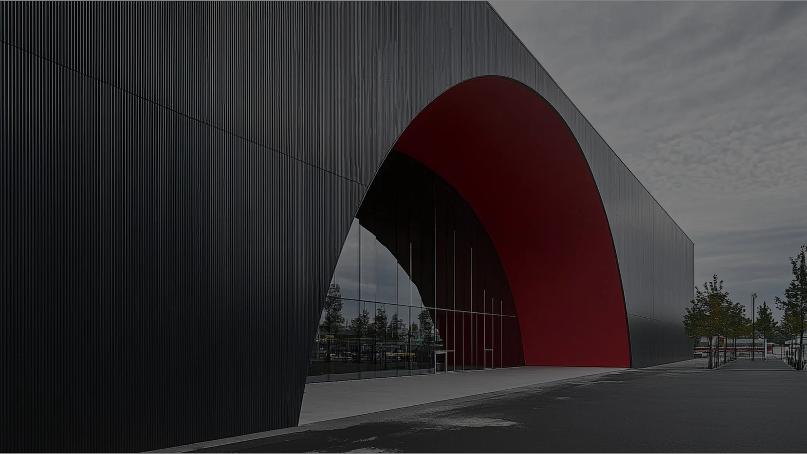 Facade of a modern, minimalist building with dark gray exterior cladding and vertical lines, featuring a large arched entrance carved into the structure, with the interior of the arch painted bright red and a large mirrored glass window at the base.