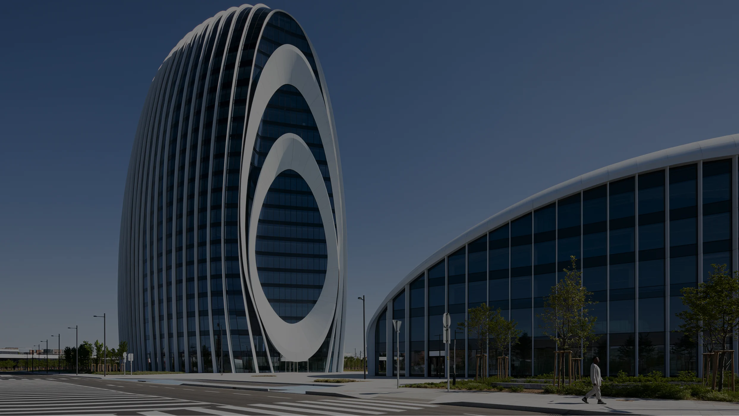A modern, ovoid-shaped skyscraper with a futuristic, white-ringed facade, flanked by a low glass building under a clear sky.