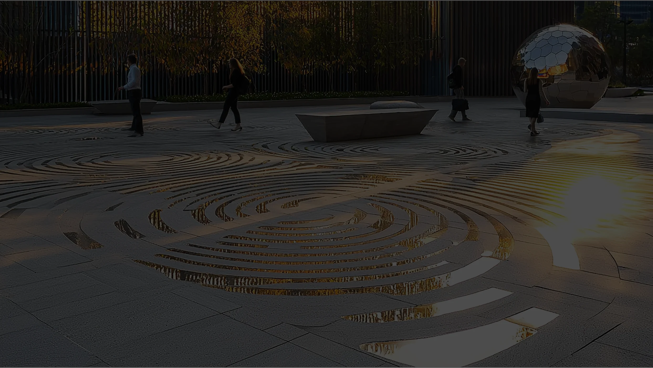 A modern pedestrian square at sunset, with people strolling among art sculptures and concentric circles of water on the pavement.