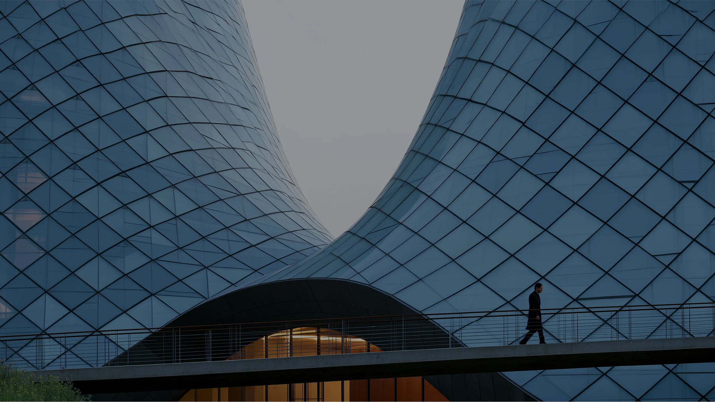 A person walks on a pedestrian walkway in front of the monumental blue glass facade of the Emporia shopping center. The architectural structure rises with two large concave, undulating shapes that curve toward each other.
