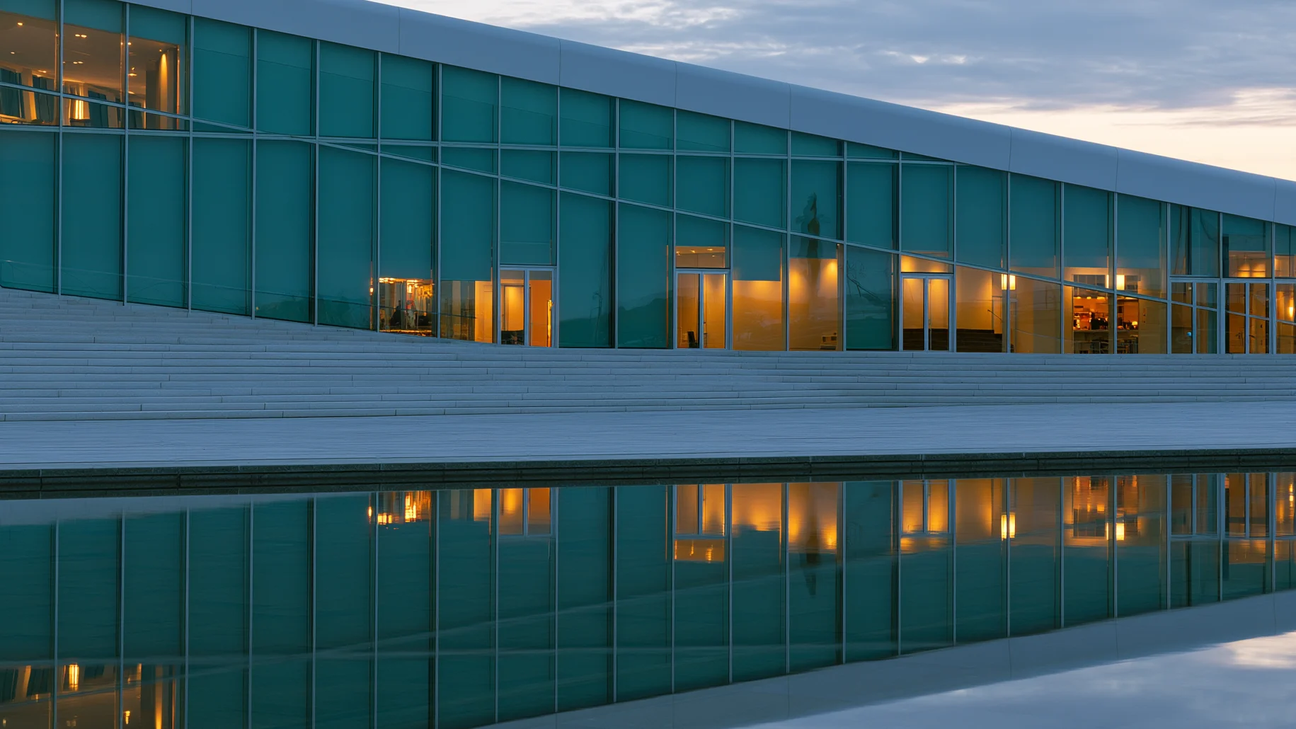 Large glass facade with warm internal lights and marble staircase, reflected on the water surface at dusk.