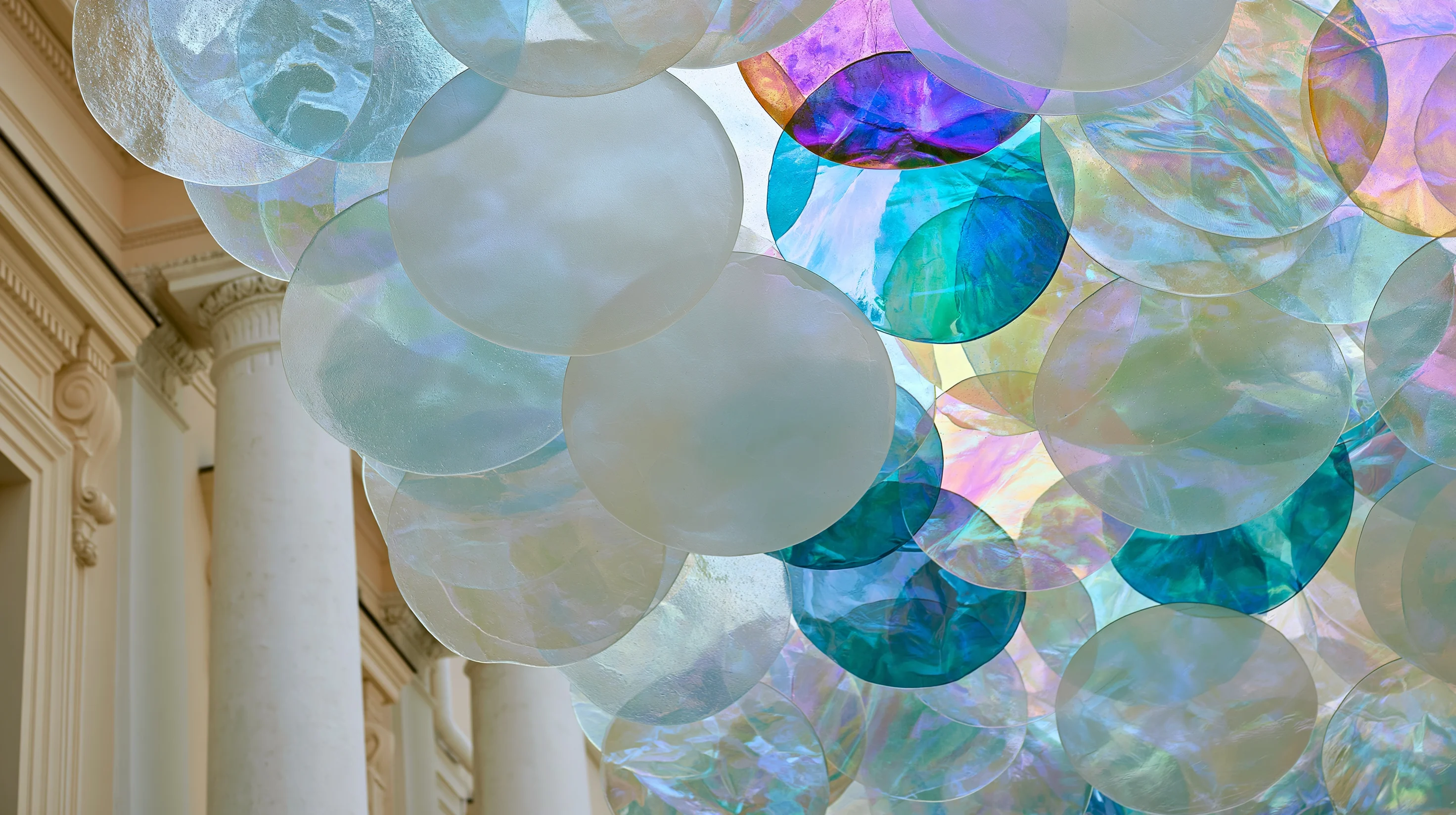 Ceiling art installation composed of transparent and iridescent discs in shades of blue, purple and pearl, suspended next to classical columns.