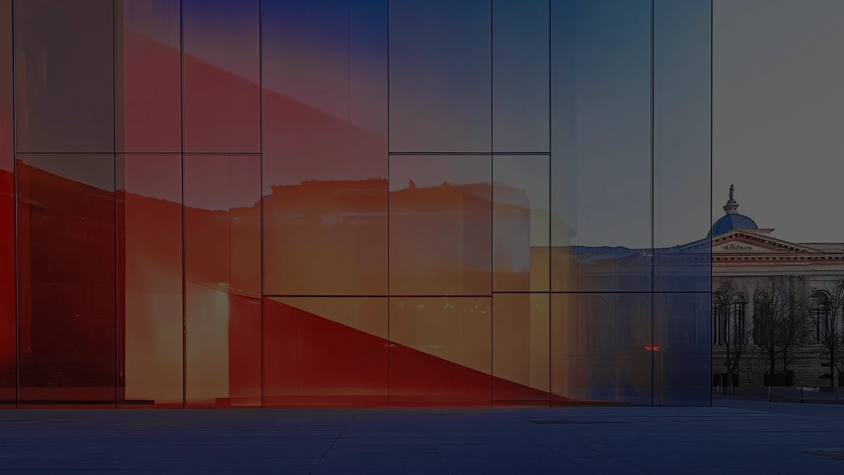 Modern geometric glass facade with chromatic shades from red to dark blue, with reflections of historic buildings and a transparent dome.