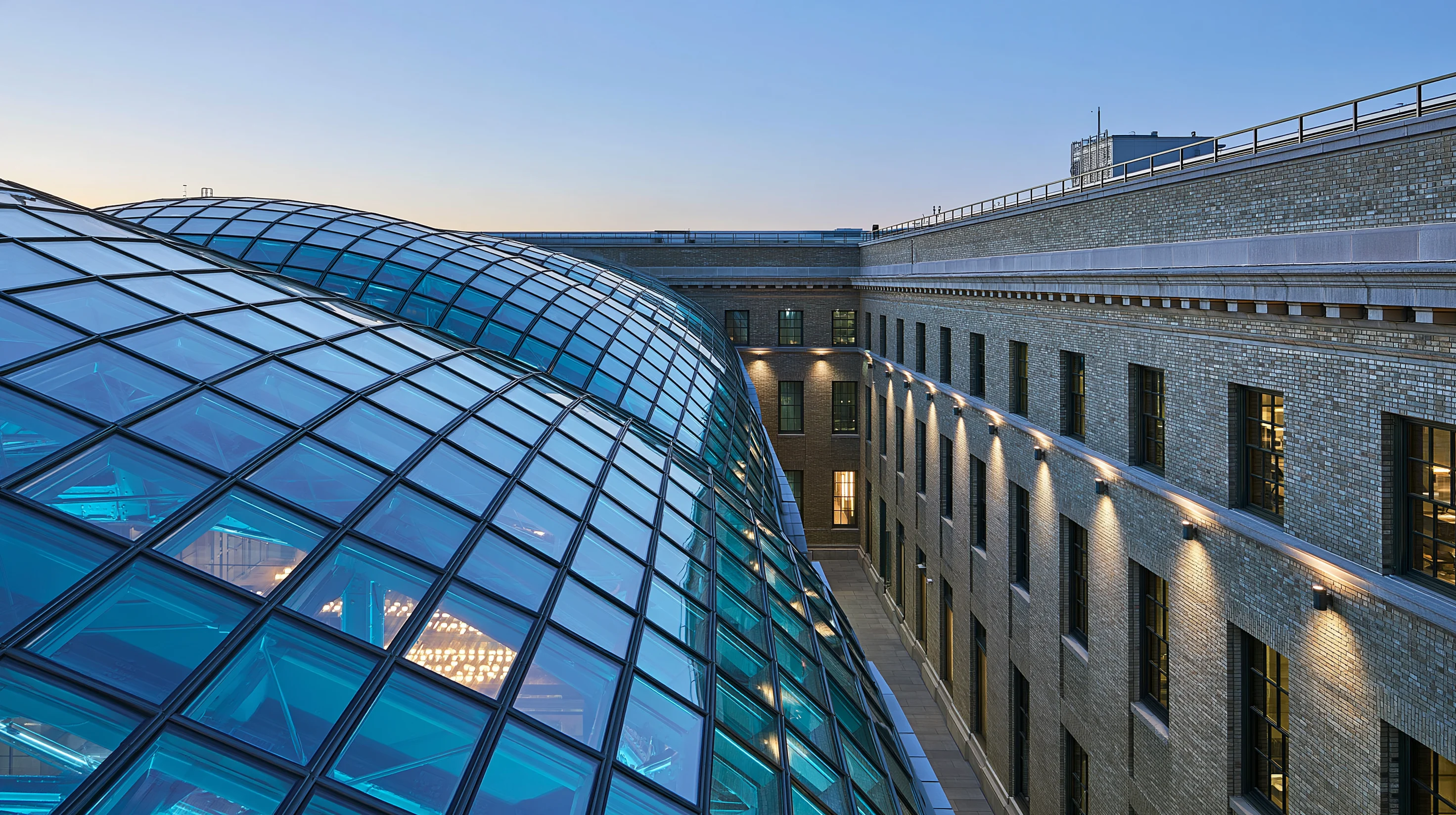 Arched roof with wavy geometric glass alongside the brick facade of a historic building illuminated at dusk.