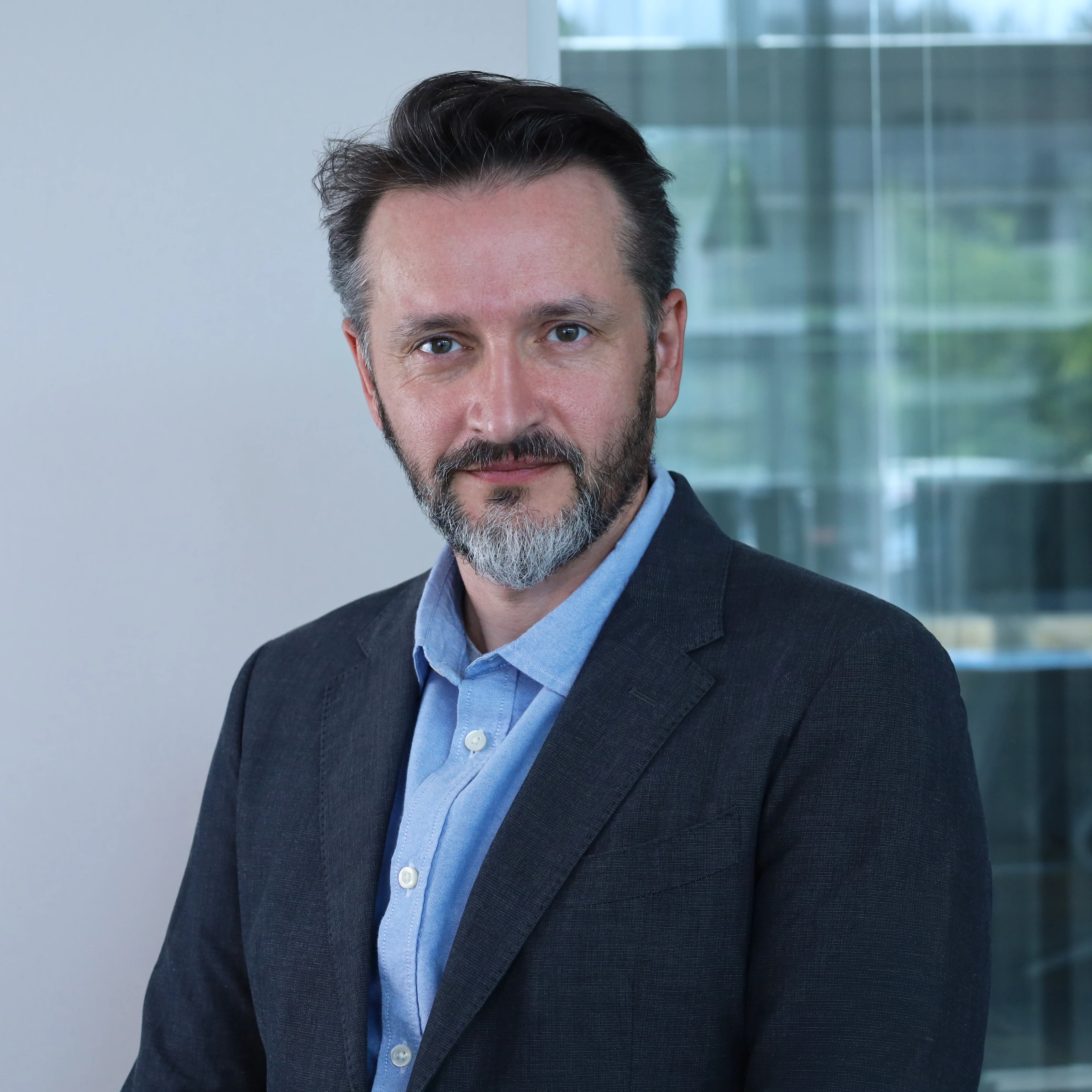 Half-length photograph of Luigi Savini, Chief DevOps Officer at Redify. A man with a graying beard, wearing a dark jacket and a blue shirt, posing inside a modern, glass-walled office.