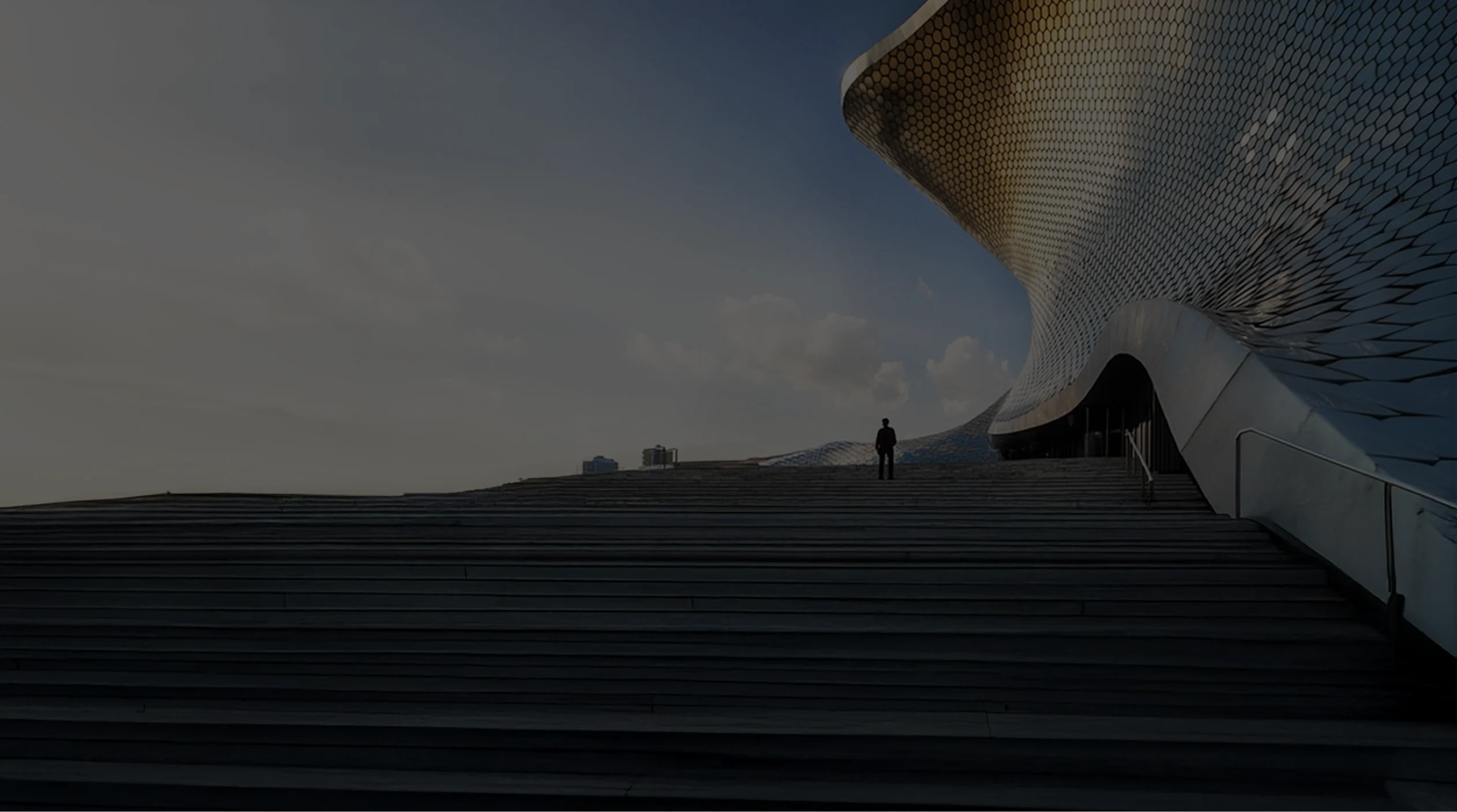 Futuristic building with curved metal facade in a hexagonal pattern and a person climbing a wide staircase under a blue sky.