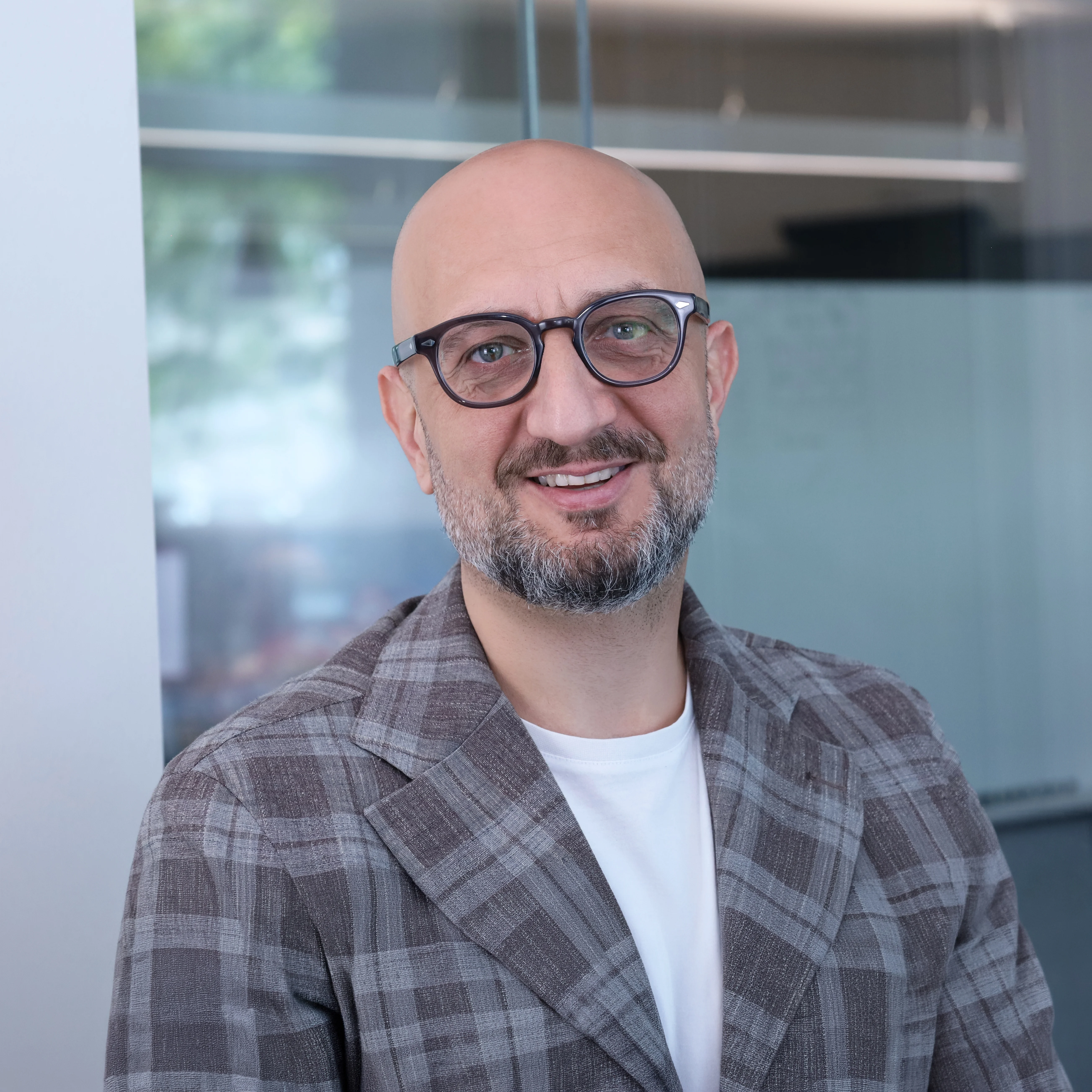 Professional portrait of Giuliano Torelli CMO from Redify: a man with glasses and a graying beard smiling at the camera. He's wearing a gray plaid jacket over a white t-shirt, against a bright, modern office backdrop.