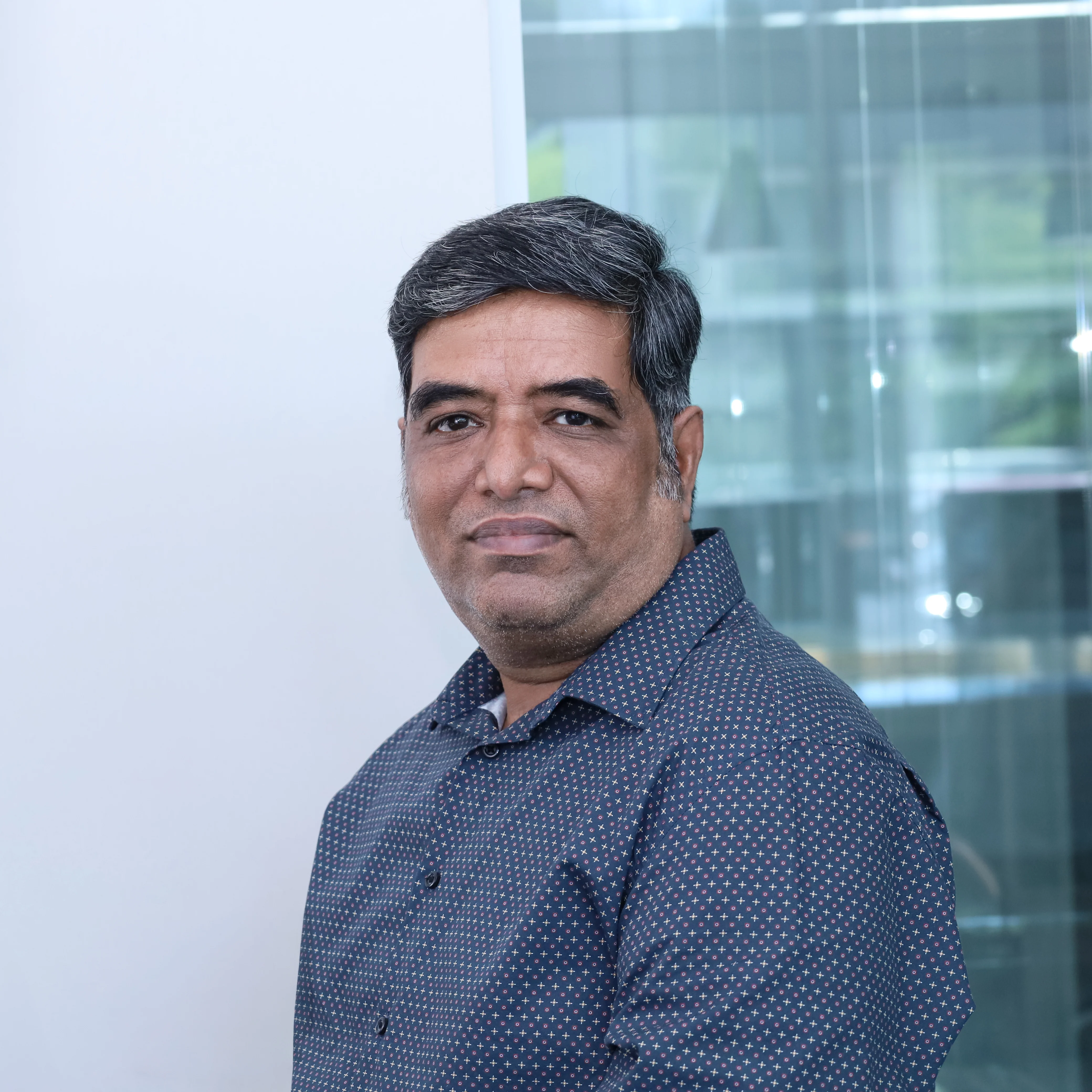 A half-length portrait of Jayaprakash Govindan, HCL, Senior Developer at Redify, wearing a dark blue geometric-patterned shirt and smiling slightly at the camera. The background is a white wall and a slightly blurred office window.