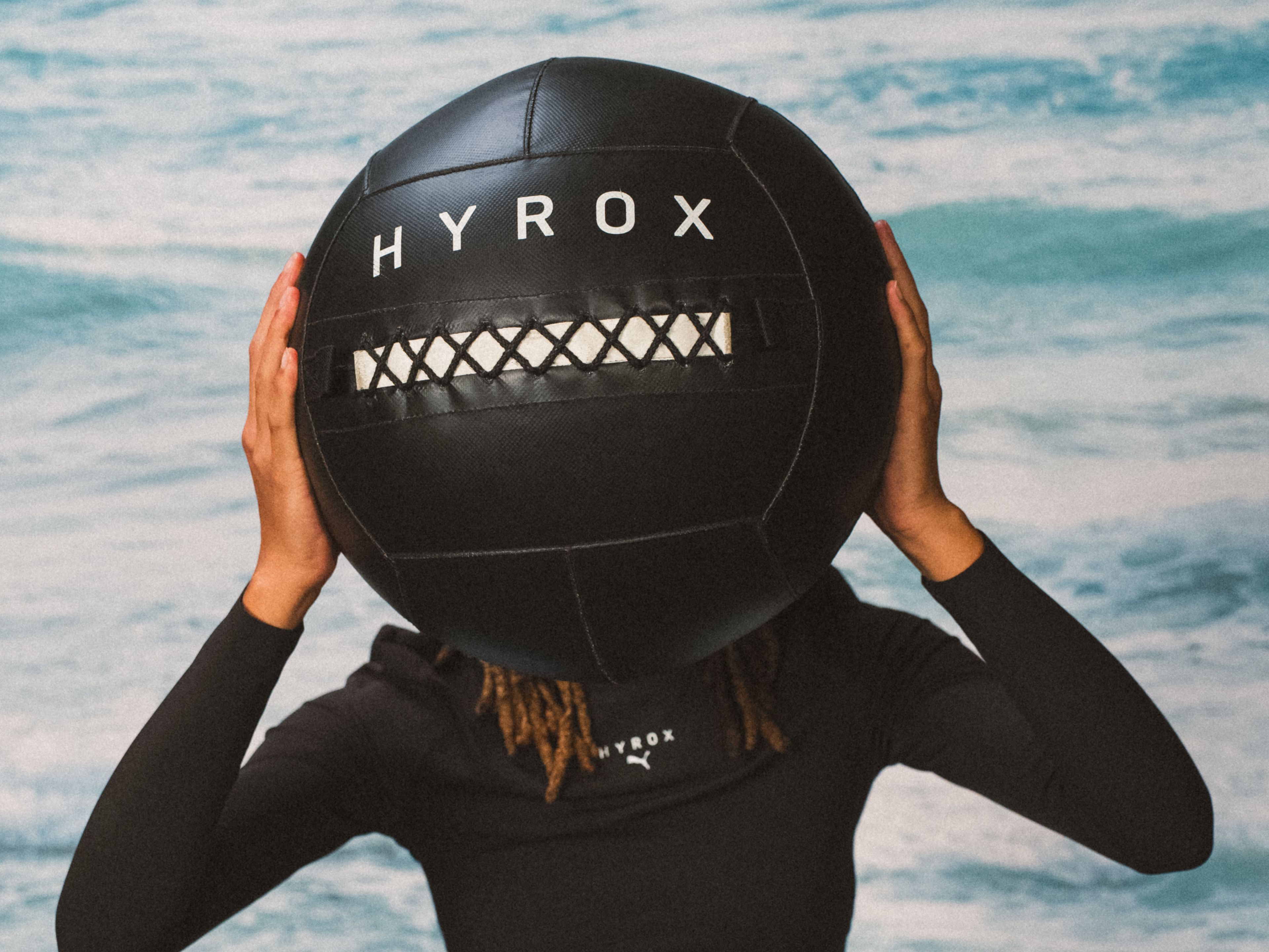 HYROC Cruise Person holding large black Hyrox ball in front of blue background.