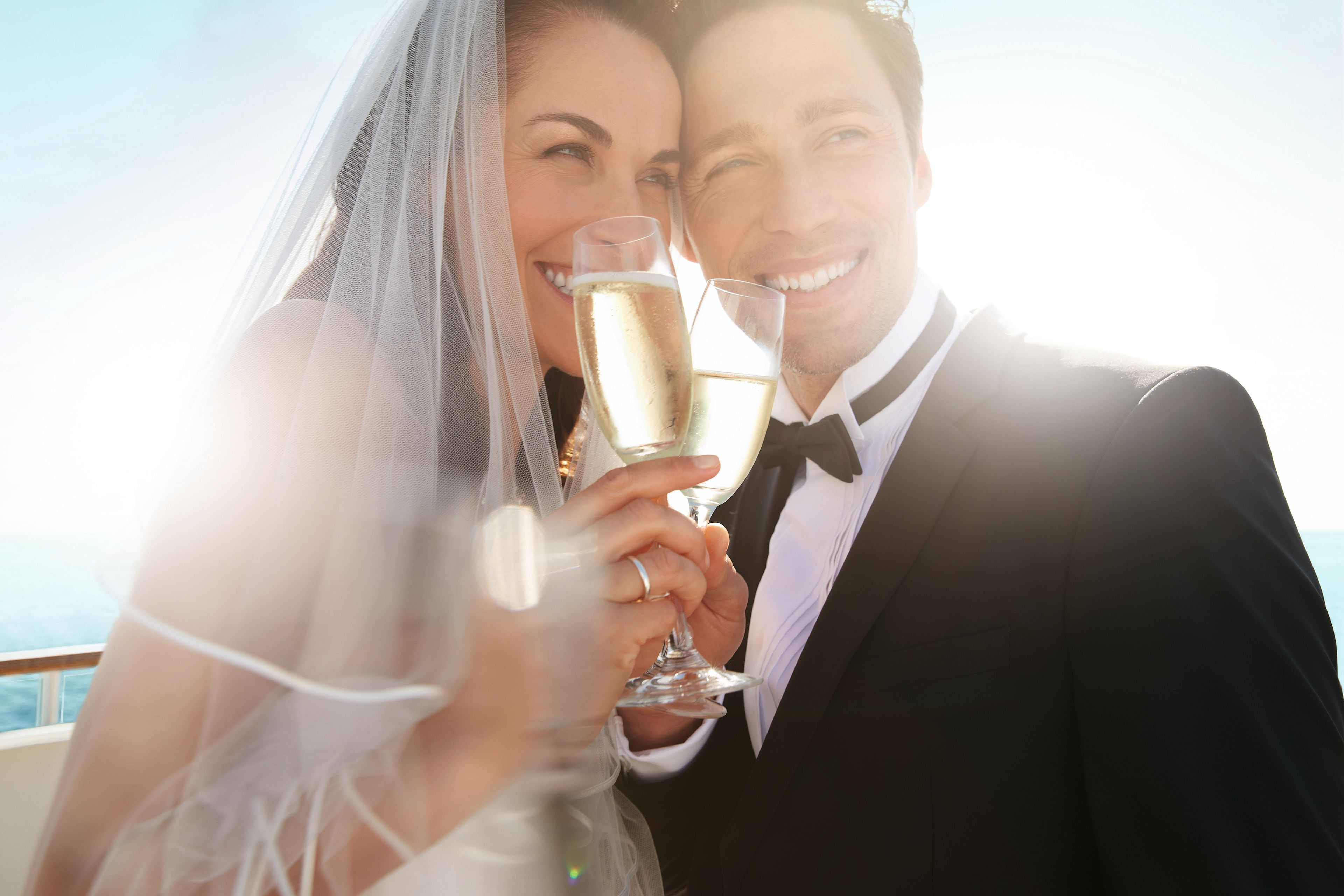Brautpaar stößt mit Champagner an, sonniger Tag, Meereshintergrund.