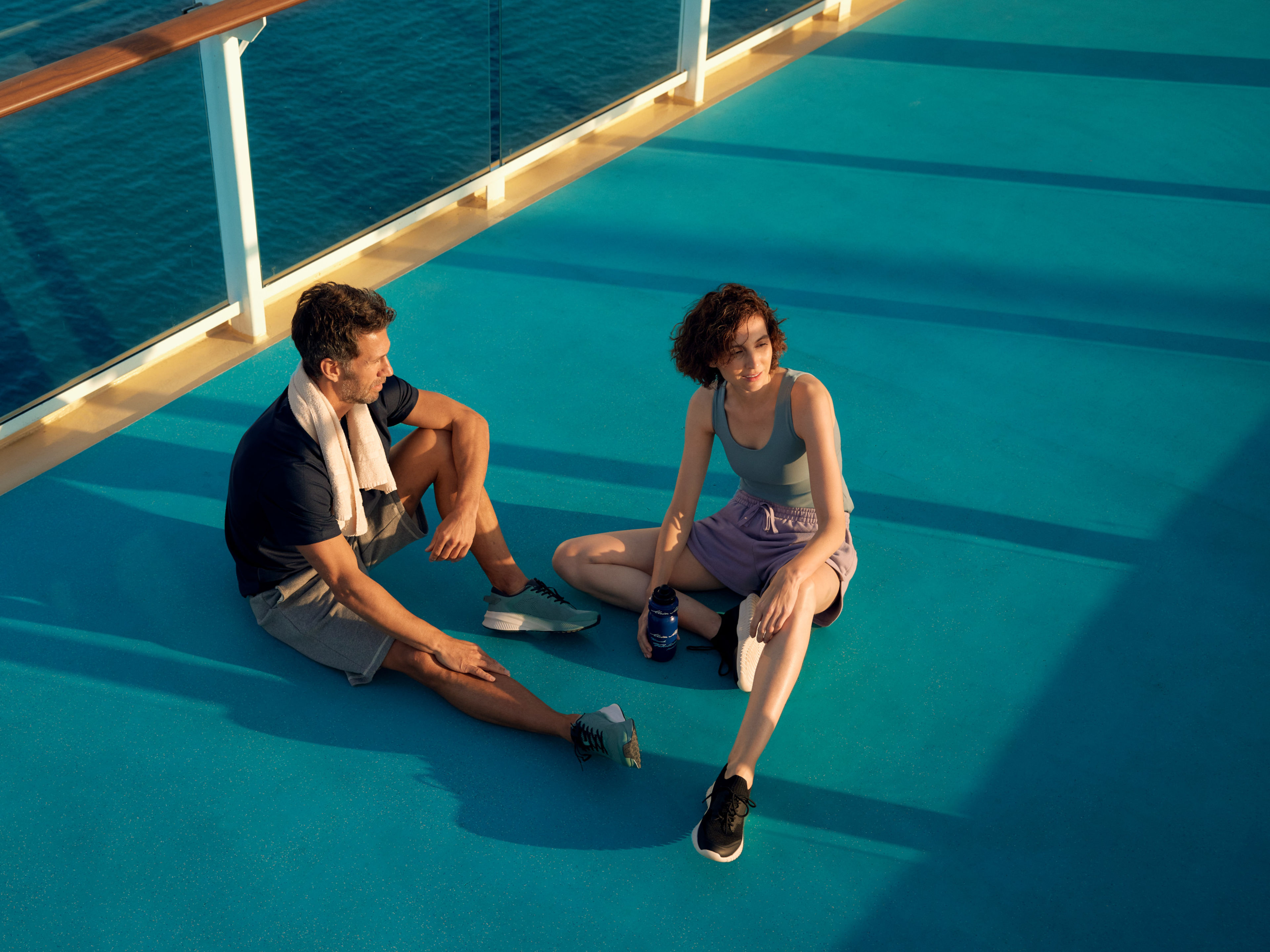 Sport auf Mein Schiff - Laufstrecke - Kampagne 2022 Zwei Personen entspannen auf dem Deck eines Kreuzfahrtschiffs bei Sonnenuntergang.
