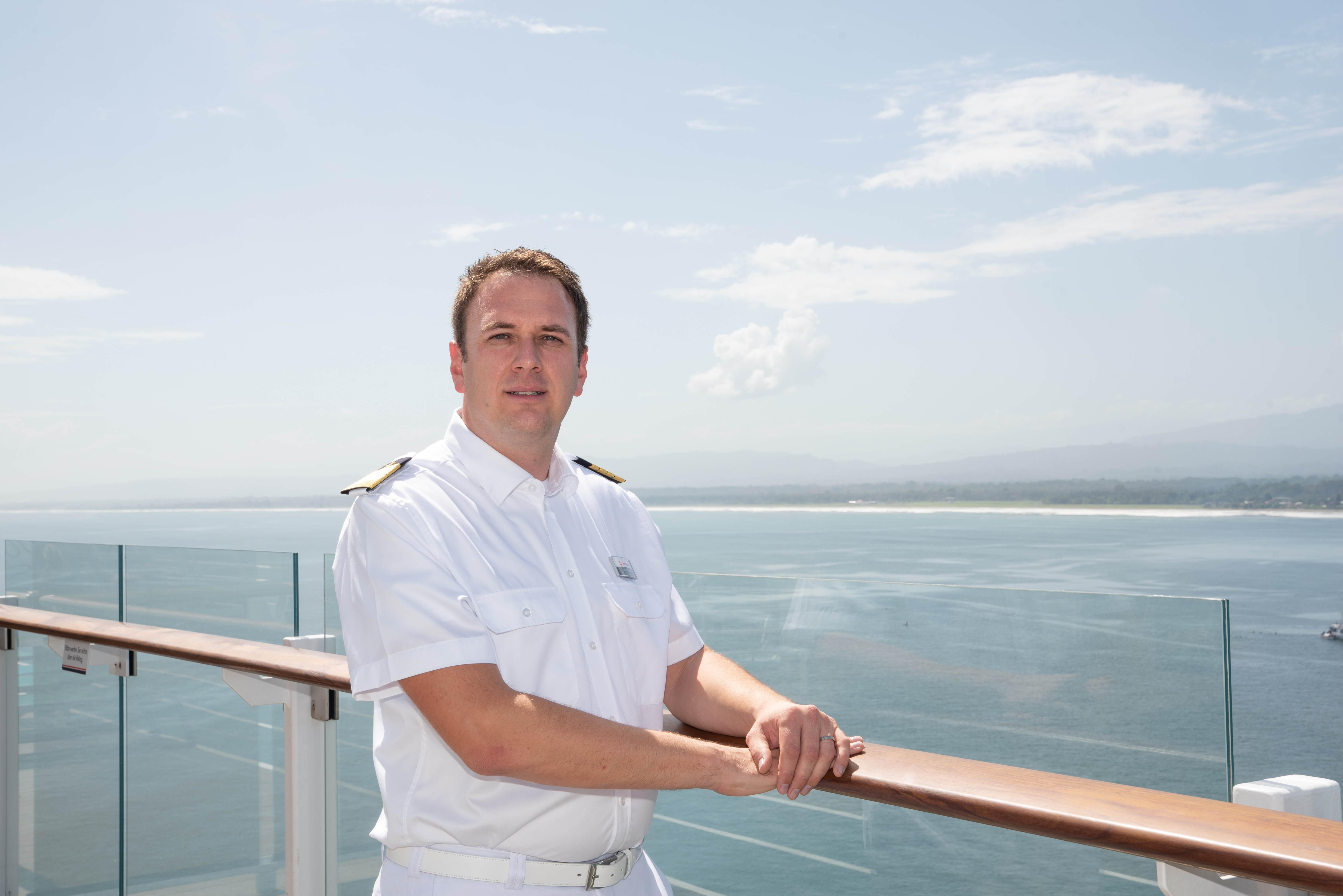 Mann in weißer Uniform auf Kreuzfahrtschiff, Meer im Hintergrund.