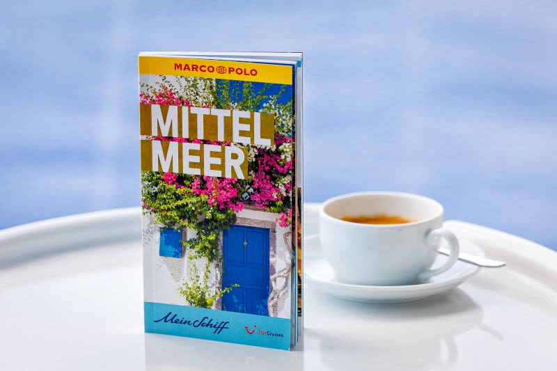Reiseführer "Mittelmeer" neben einer Tasse Kaffee auf weißem Tisch.