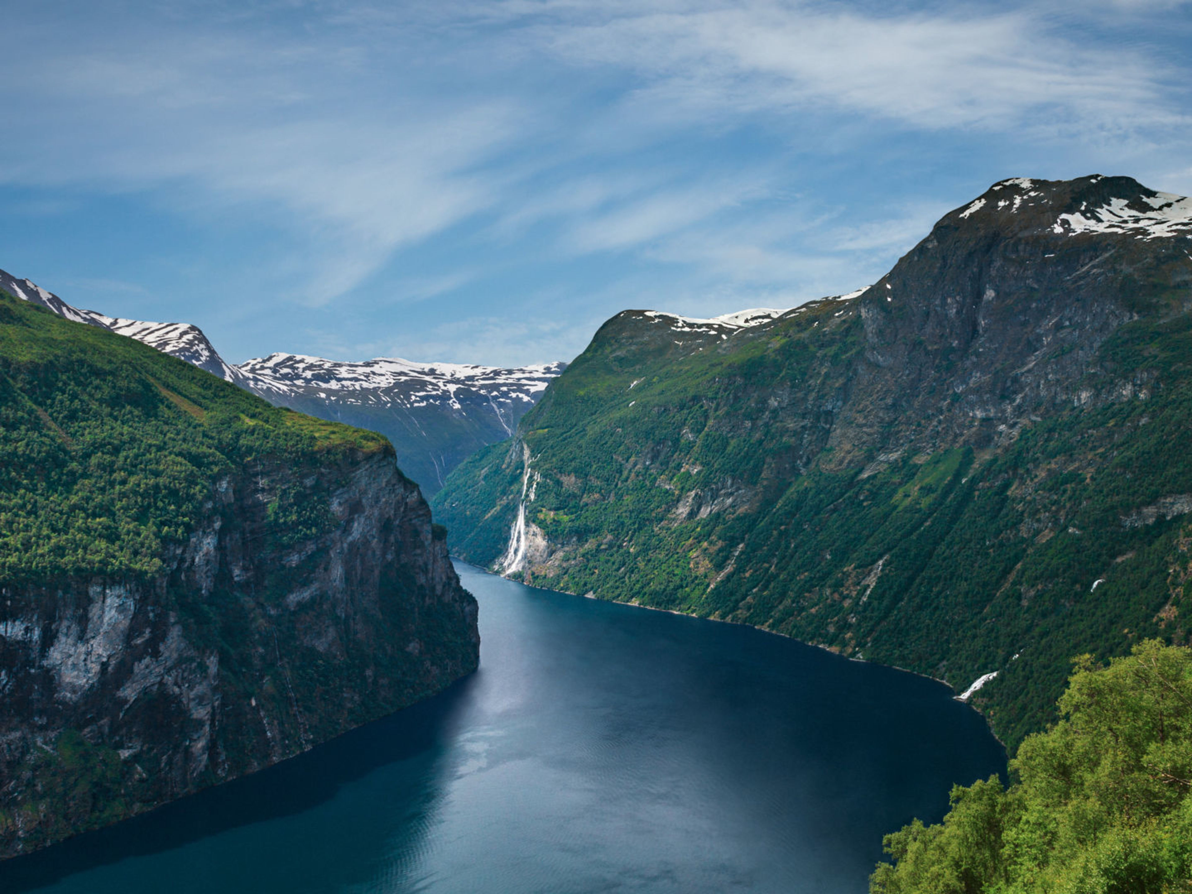 Geiranger Fjord Norwegischer Fjord mit Wasserfall, umgeben von grünen Bergen und blauem Himmel.