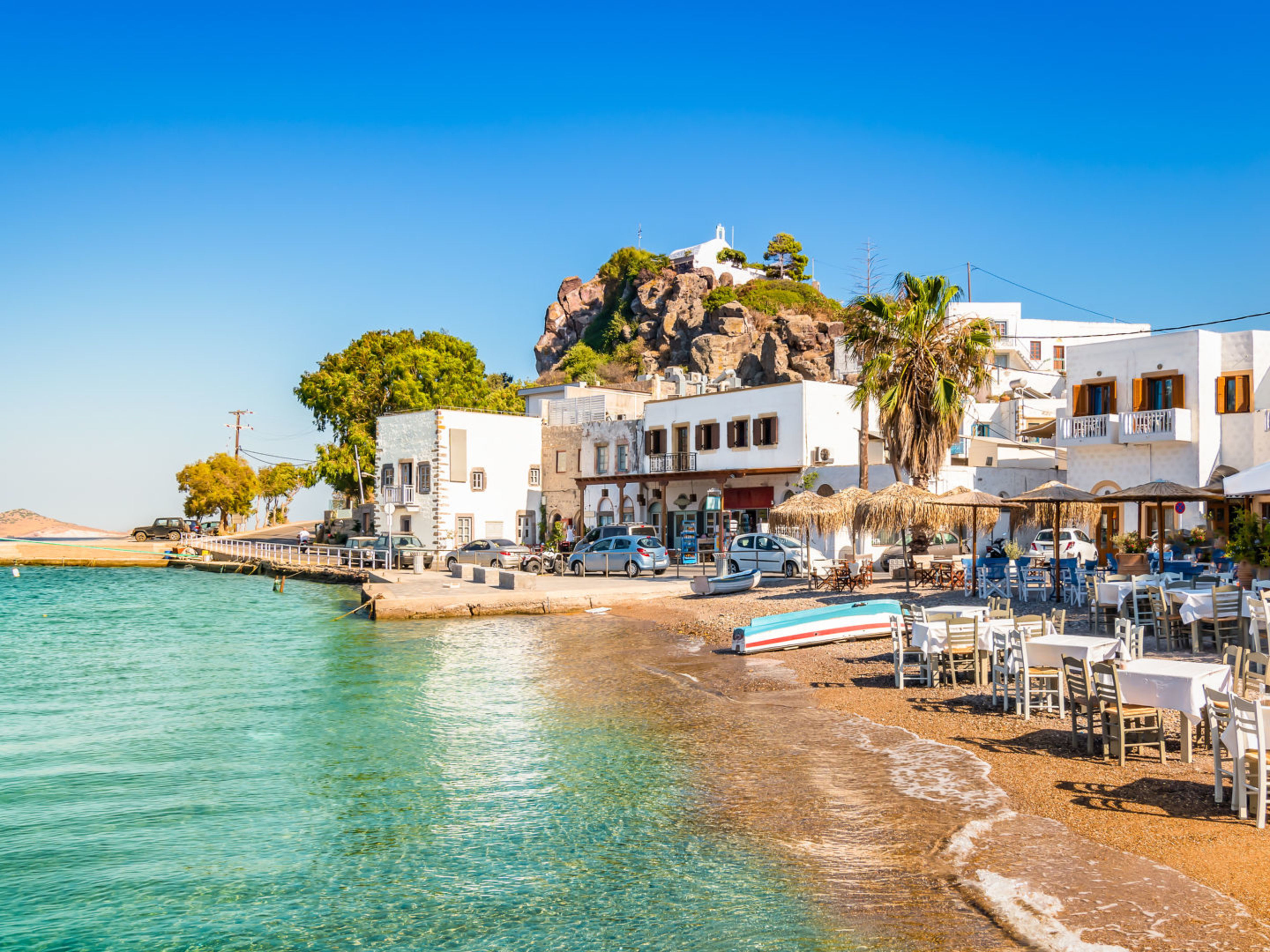 Destinationsmotiv, Patmos, Griechenland Strandpromenade mit weißen Gebäuden und Tischen, blaues Meer, blauer Himmel.