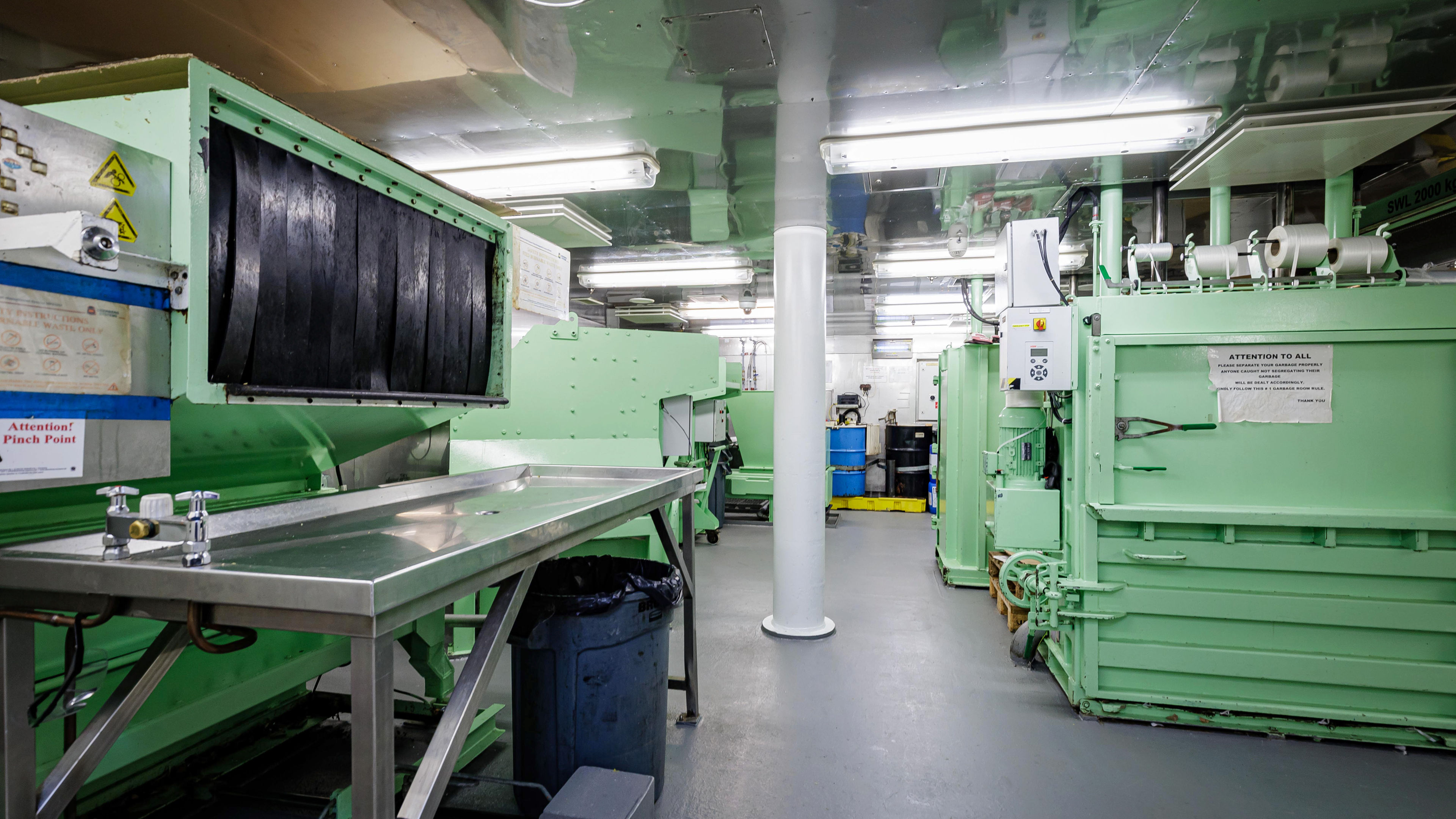Grüner Maschinenraum mit Metalltischen und blauen Fässern.