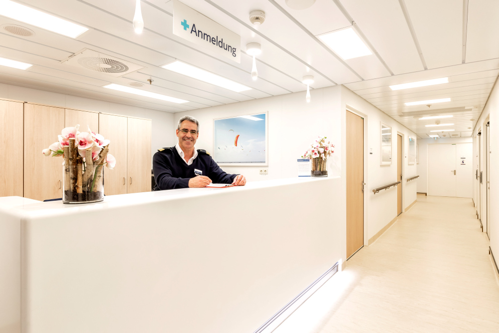 Mann an Rezeption, lächelnd, weiße Wände, Blumen, Anmeldungsschild.