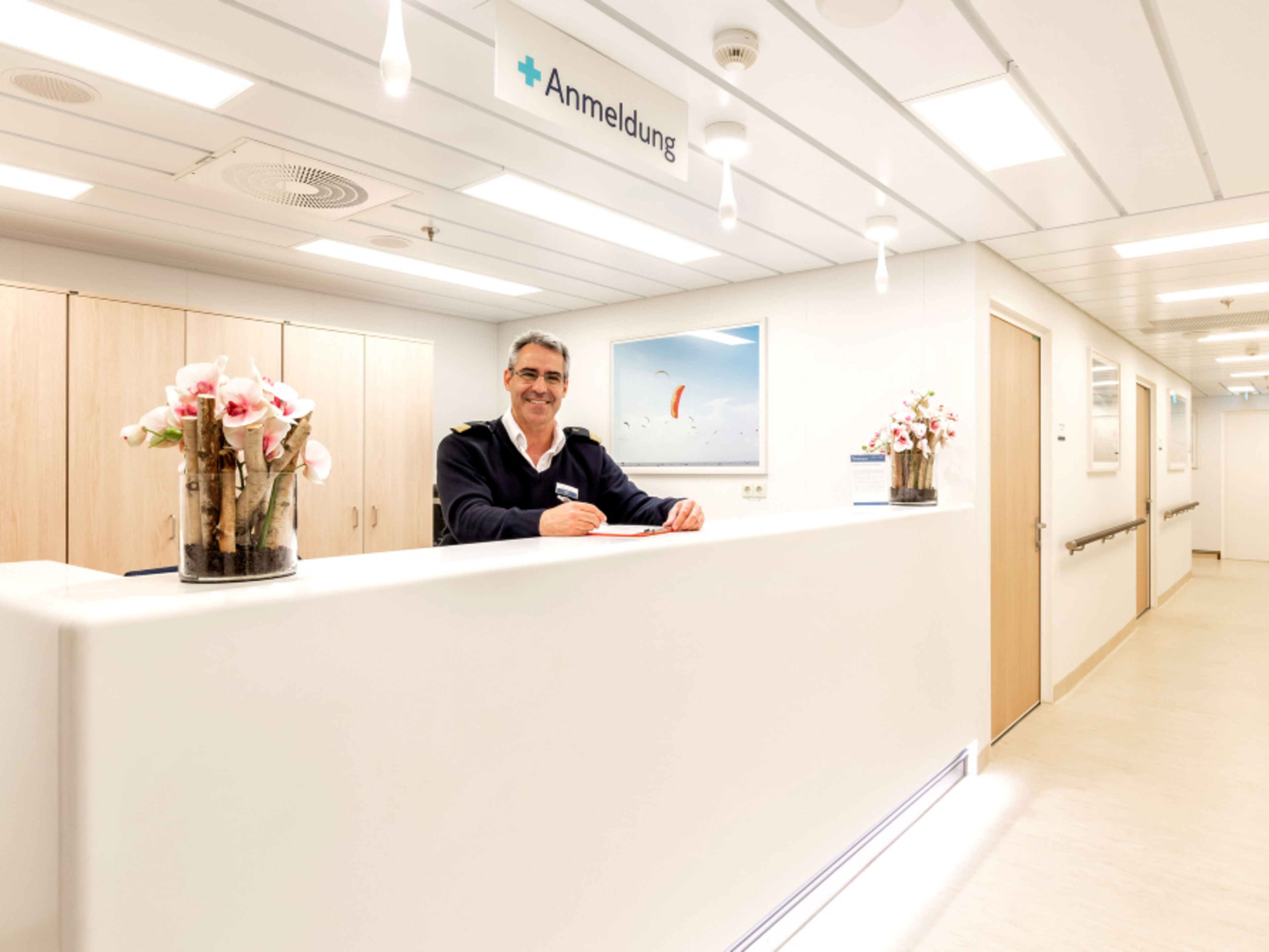 Bordhospital Mein Schiff 1 Mann an Rezeption, lächelnd, weiße Wände, Blumen, Anmeldungsschild.