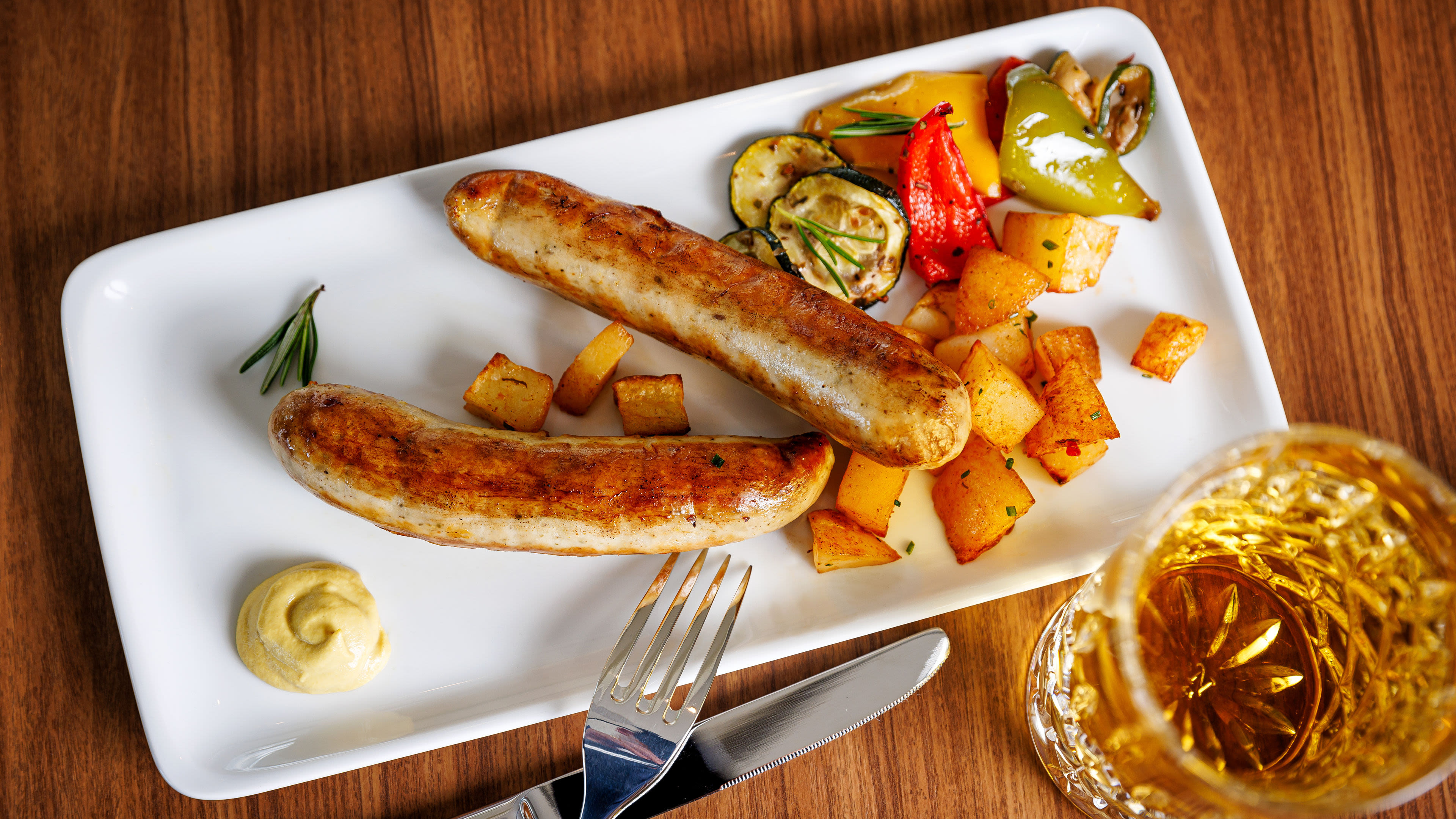 Bratwürste mit Kartoffeln und Gemüse auf weißem Teller, Glas Bier.