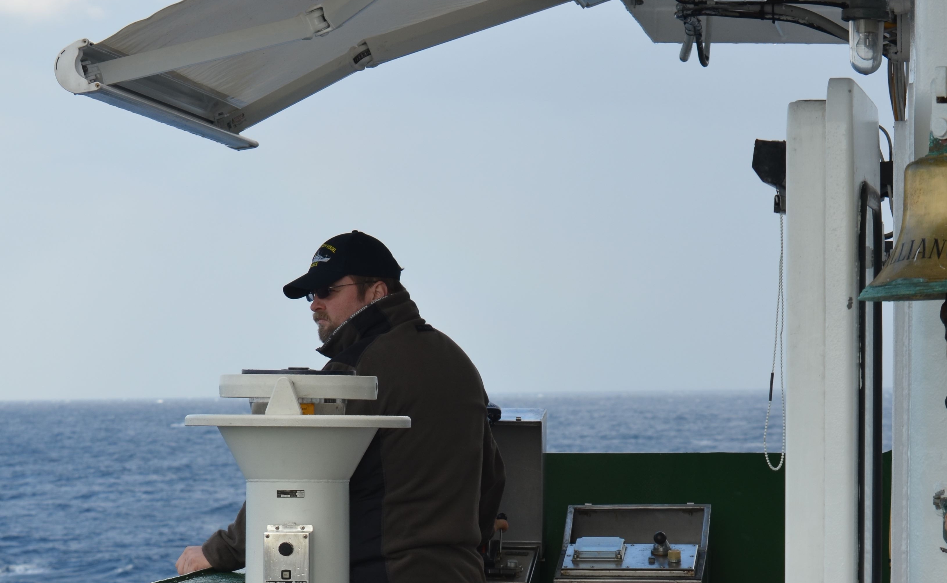 Mann mit Mütze auf Schiff, Meer im Hintergrund.