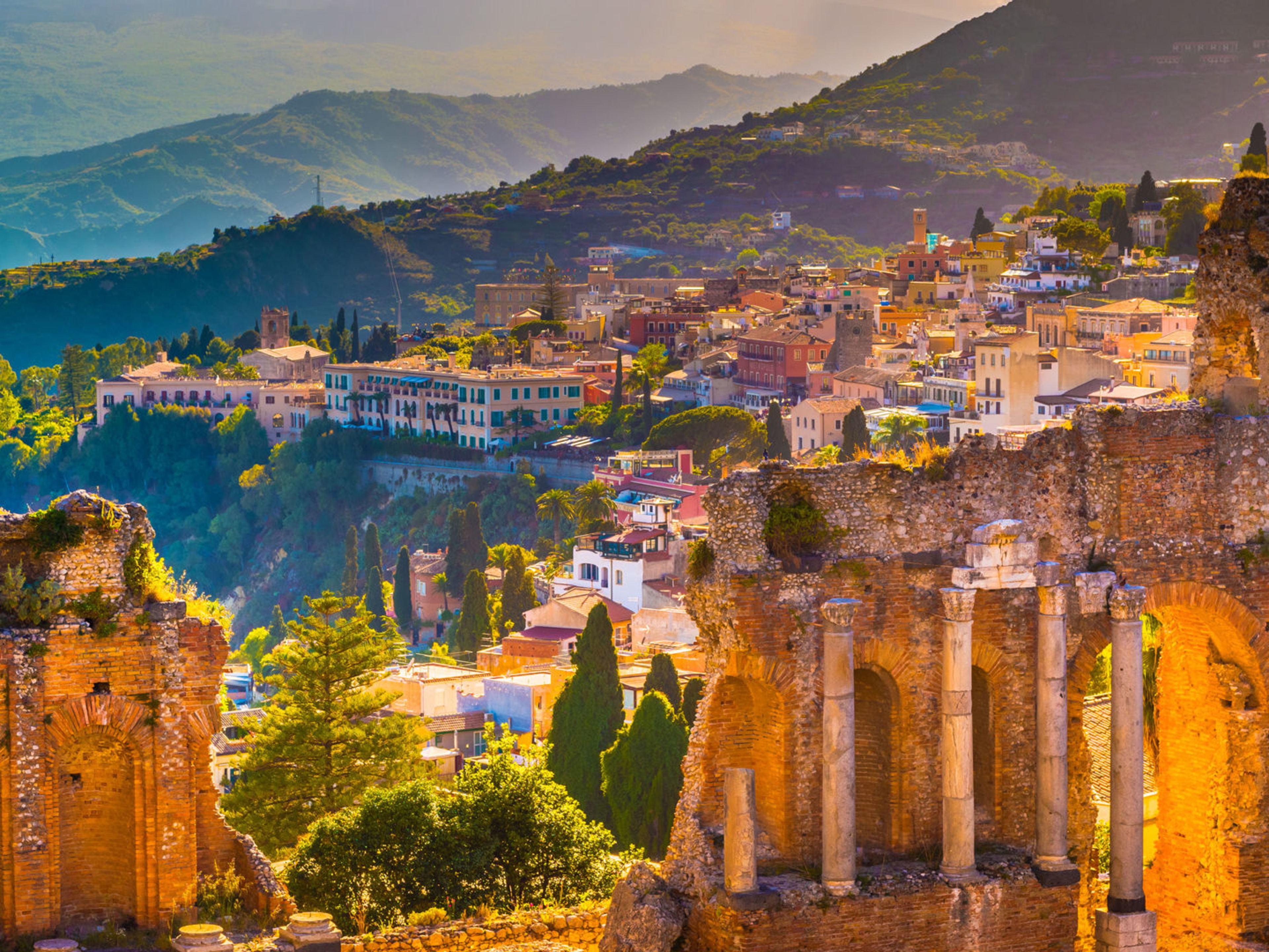 Sizilien, Italien Antike Ruinen in Taormina, Sizilien, mit bunten Häusern und grünen Hügeln im Hintergrund.