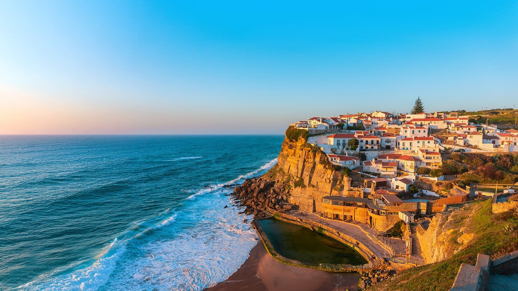 Küstenstadt auf Klippe bei Sonnenuntergang, blaues Meer, bunte Häuser.
