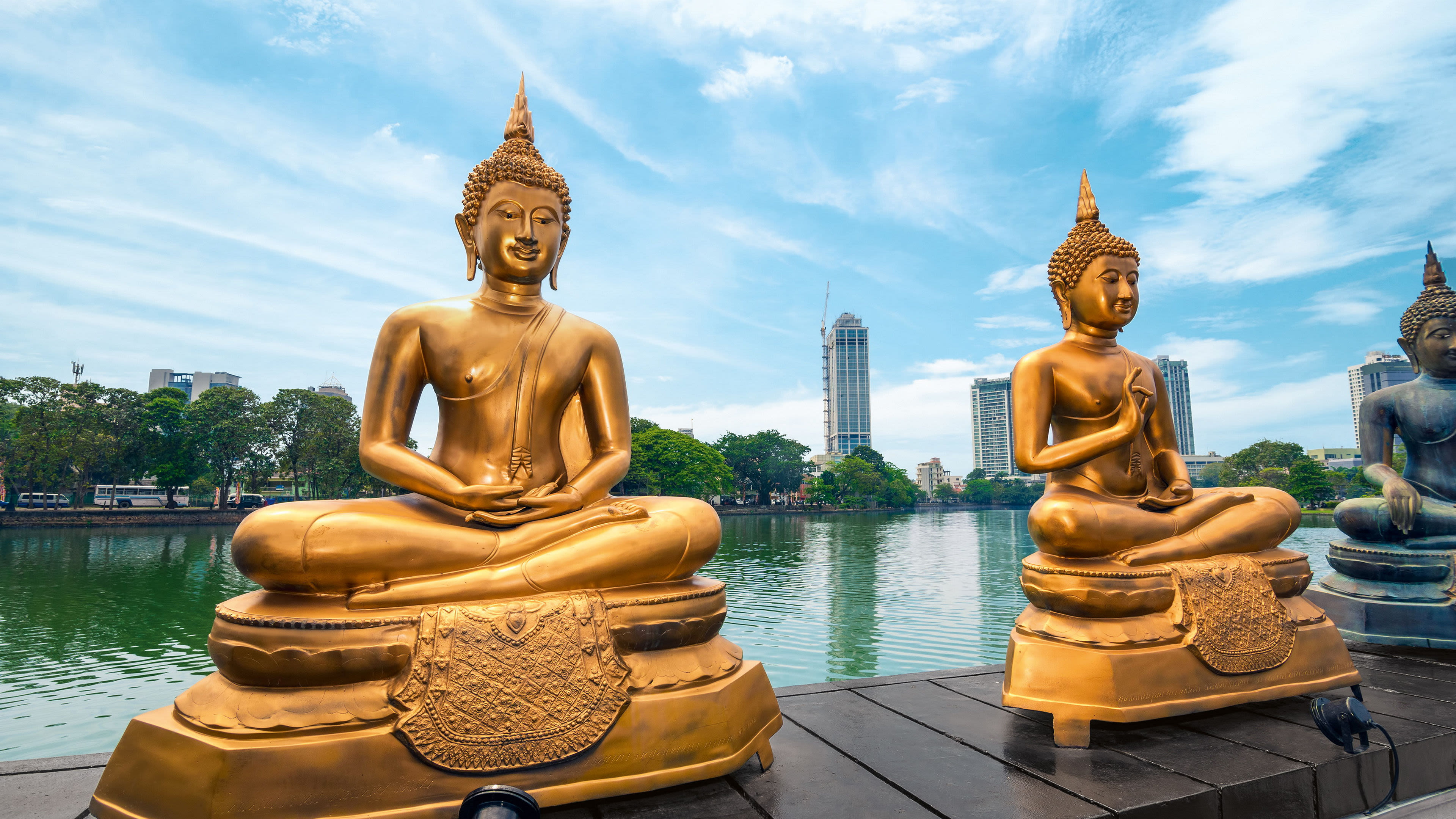 Goldene Buddha-Statuen am Wasser, Stadt im Hintergrund, blauer Himmel.