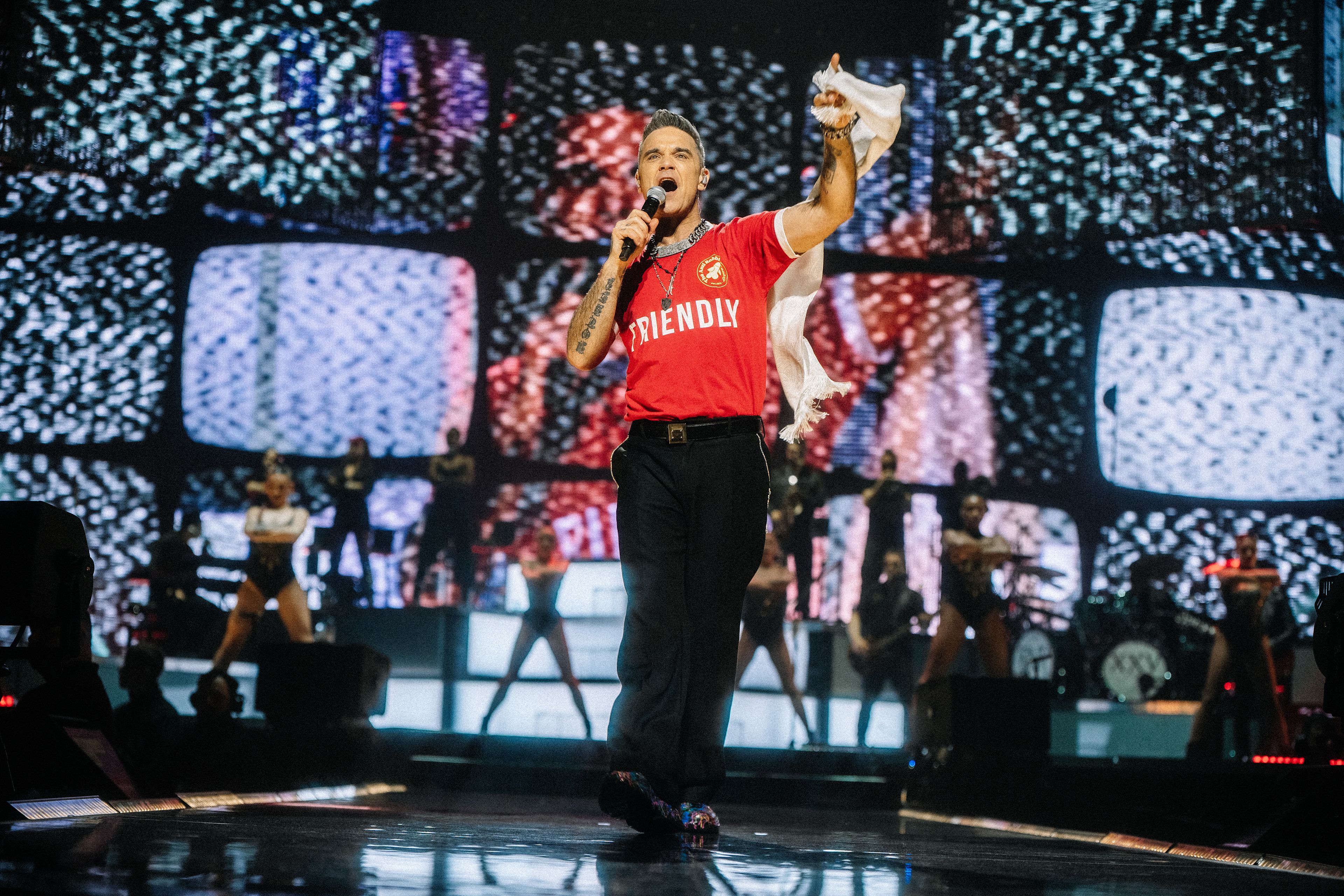 Sänger in rotem Shirt auf Bühne mit Tänzern und Lichtern.