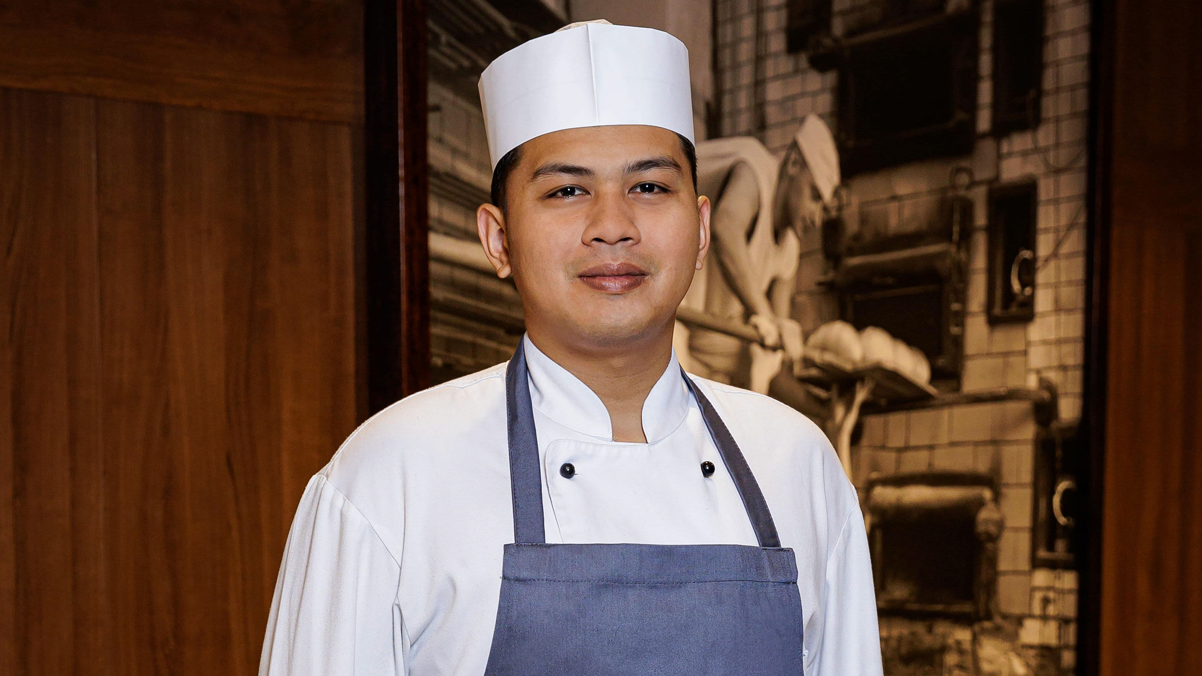 Koch in weißer Uniform und Schürze vor Holzwand.