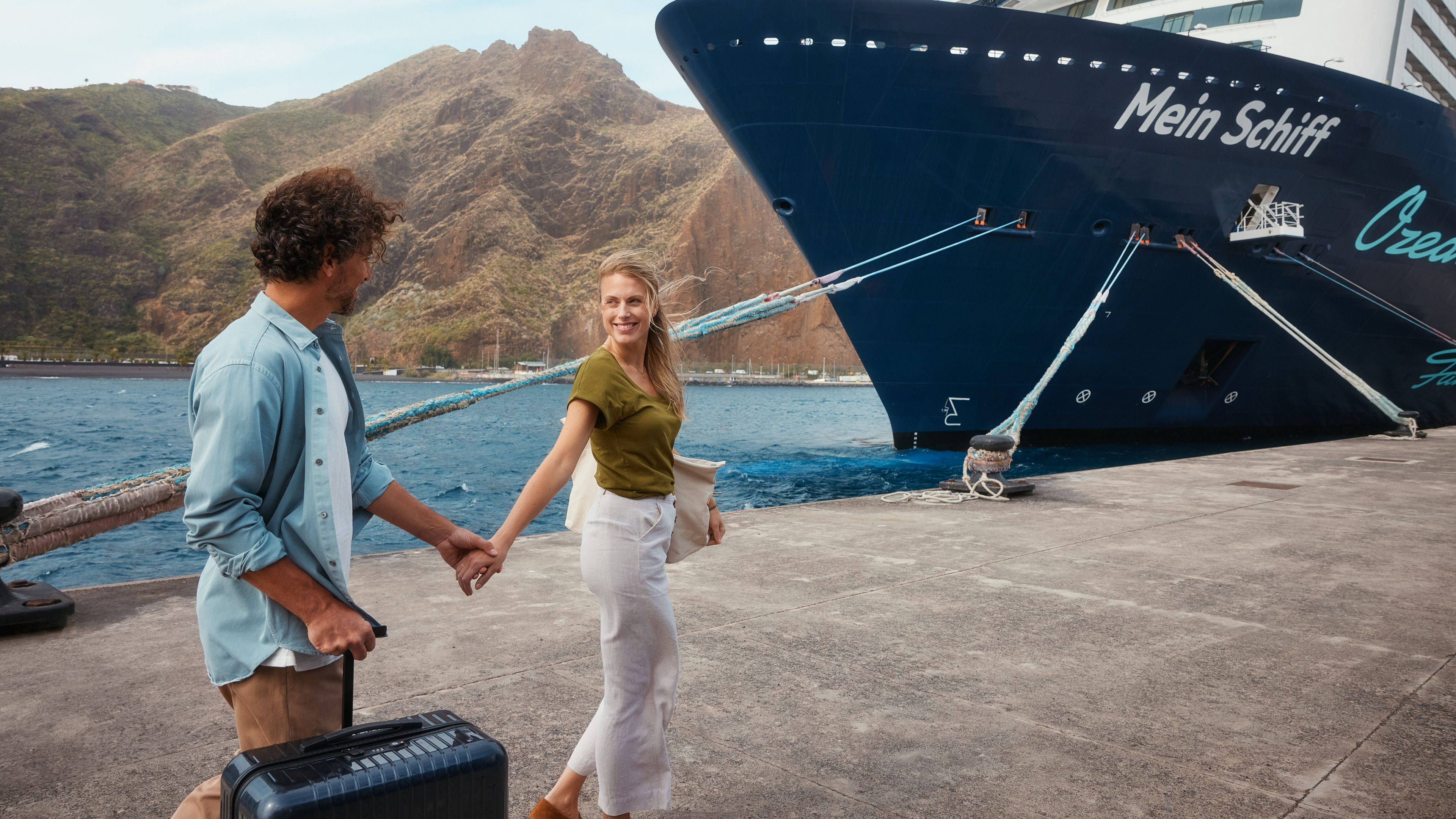 Paar am Hafen vor Kreuzfahrtschiff der Mein Schiff® Flotte, Berge im Hintergrund.