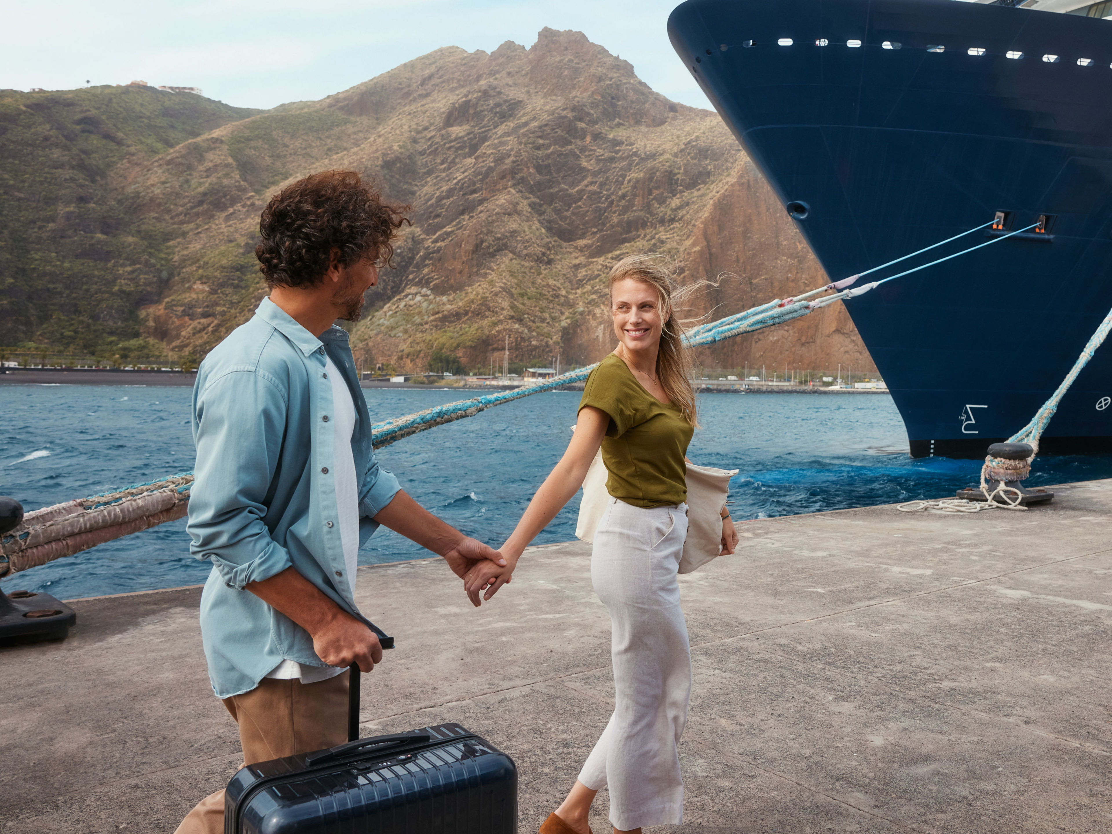 Paar an der Pier vorm Schiff Paar am Hafen vor Kreuzfahrtschiff der Mein Schiff® Flotte, Berge im Hintergrund.