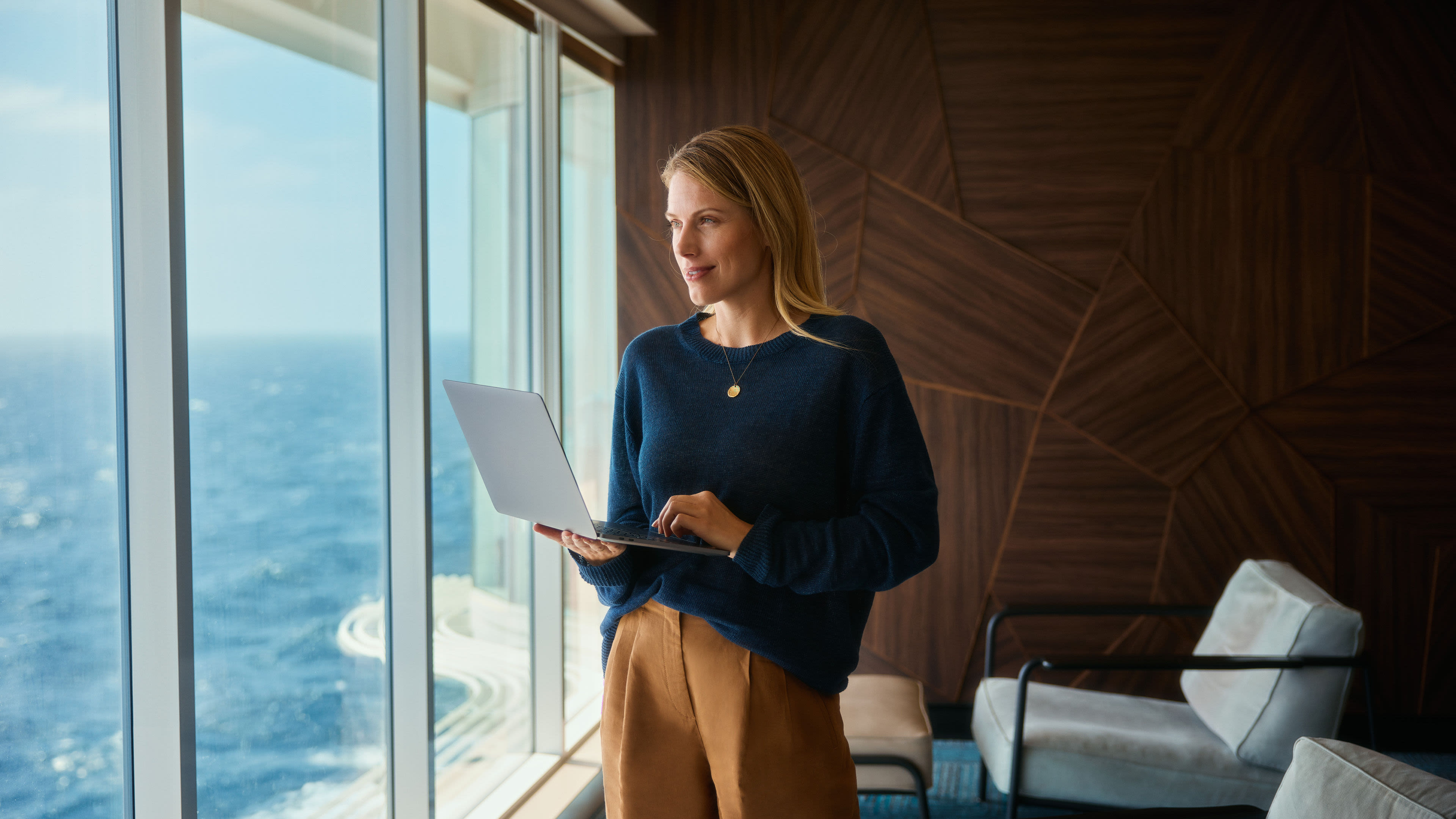 Frau mit Laptop, blickt aus Fenster auf Meer.