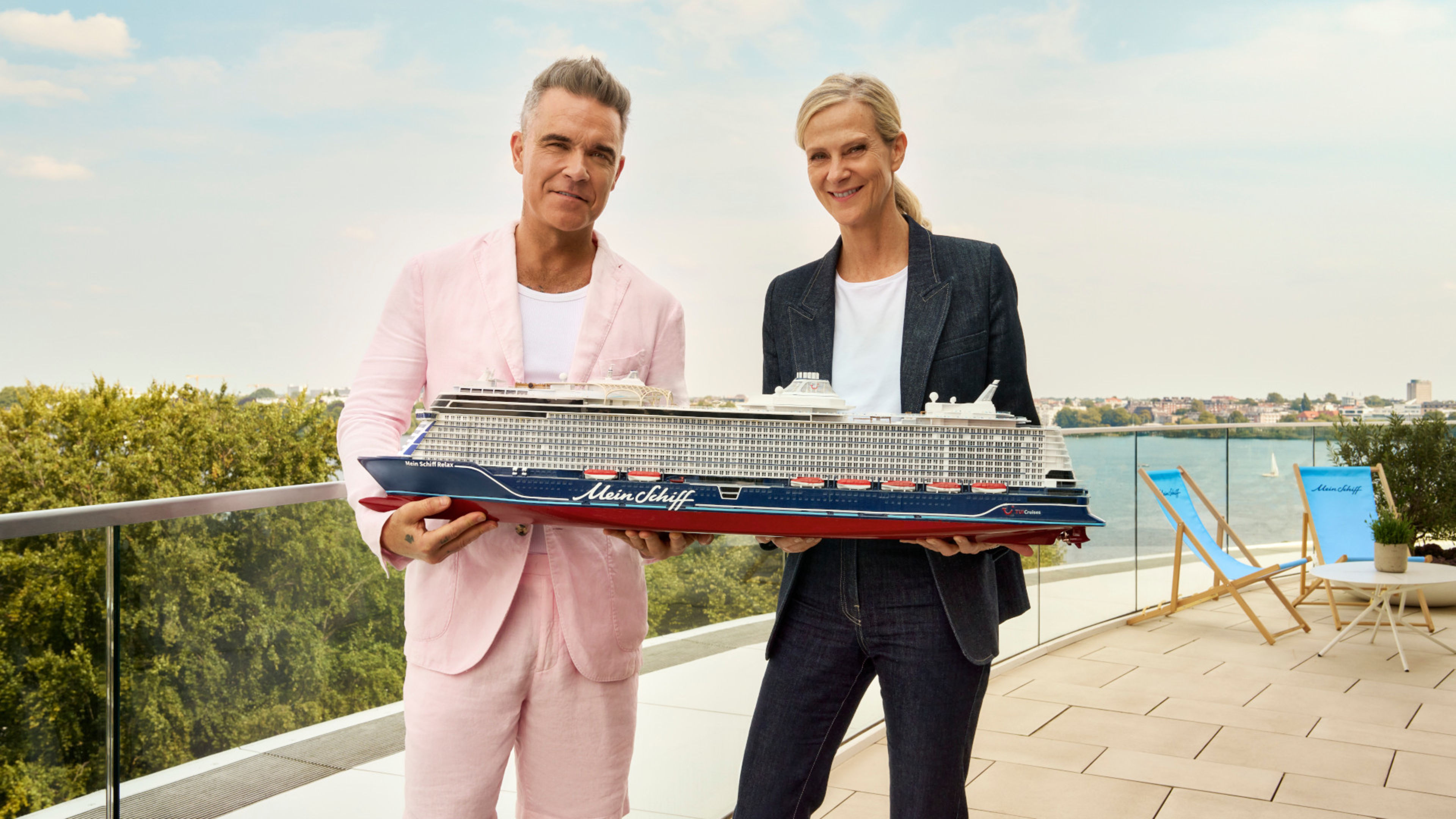 Zwei Personen halten Modell eines Kreuzfahrtschiffs der Mein Schiff® Flotte auf Terrasse mit Seeblick. Zwei Personen halten Modell eines Kreuzfahrtschiffs der Mein Schiff® Flotte auf Terrasse mit Seeblick.