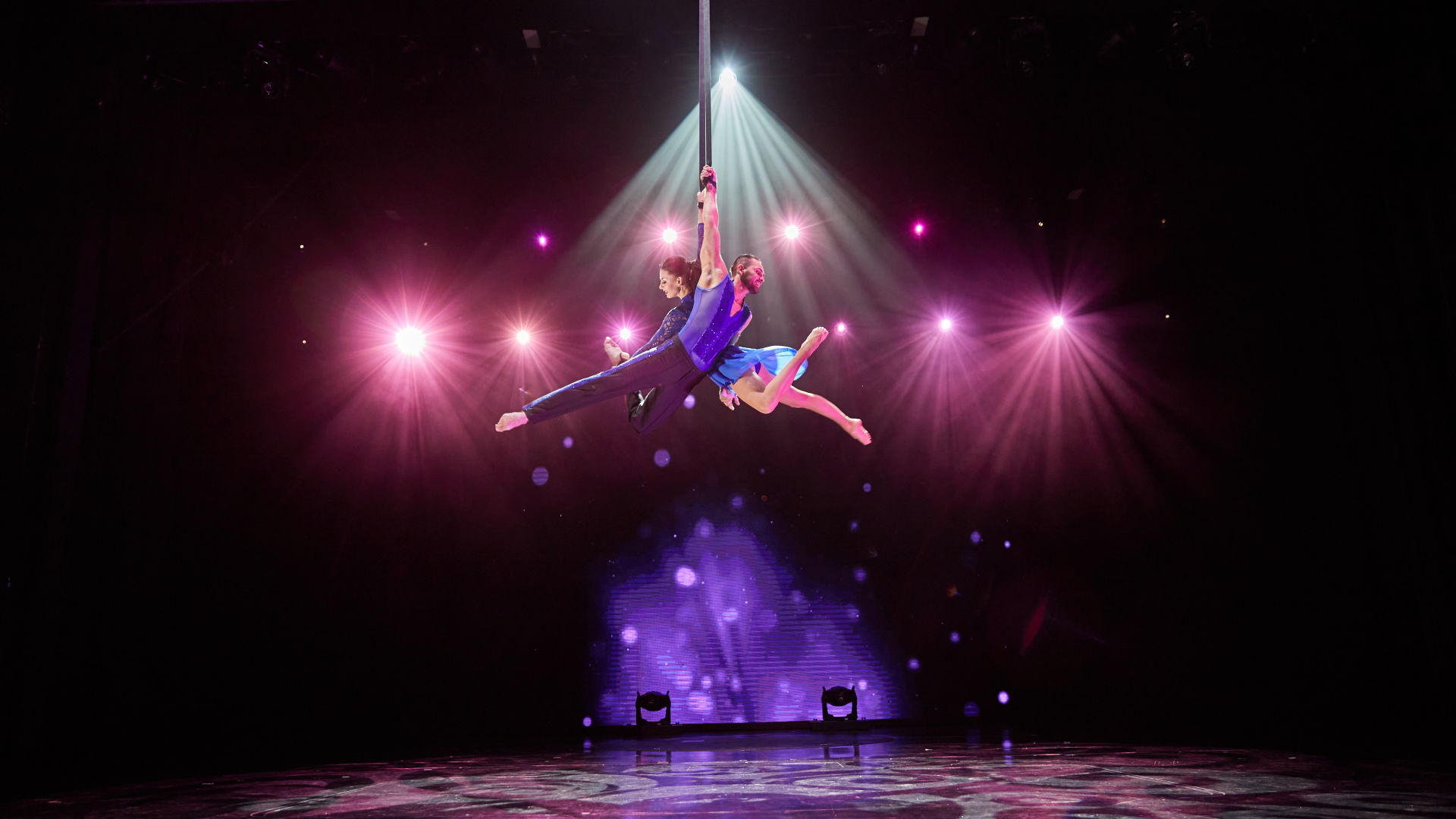 Zwei Akrobaten in blauen Kostümen schweben unter bunten Lichtern.