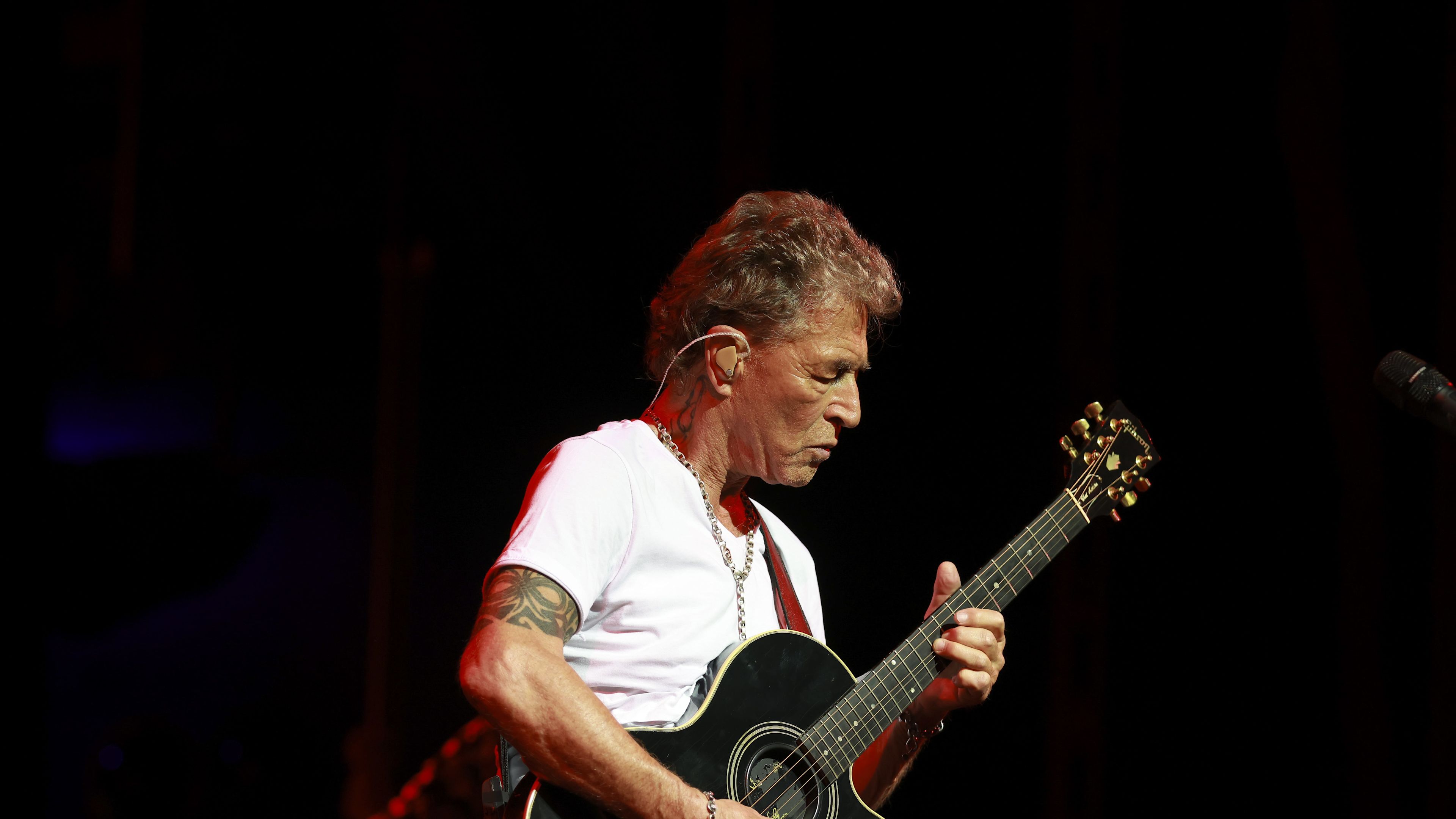 Peter Maffay Mann spielt Gitarre auf Bühne, trägt weißes T-Shirt.