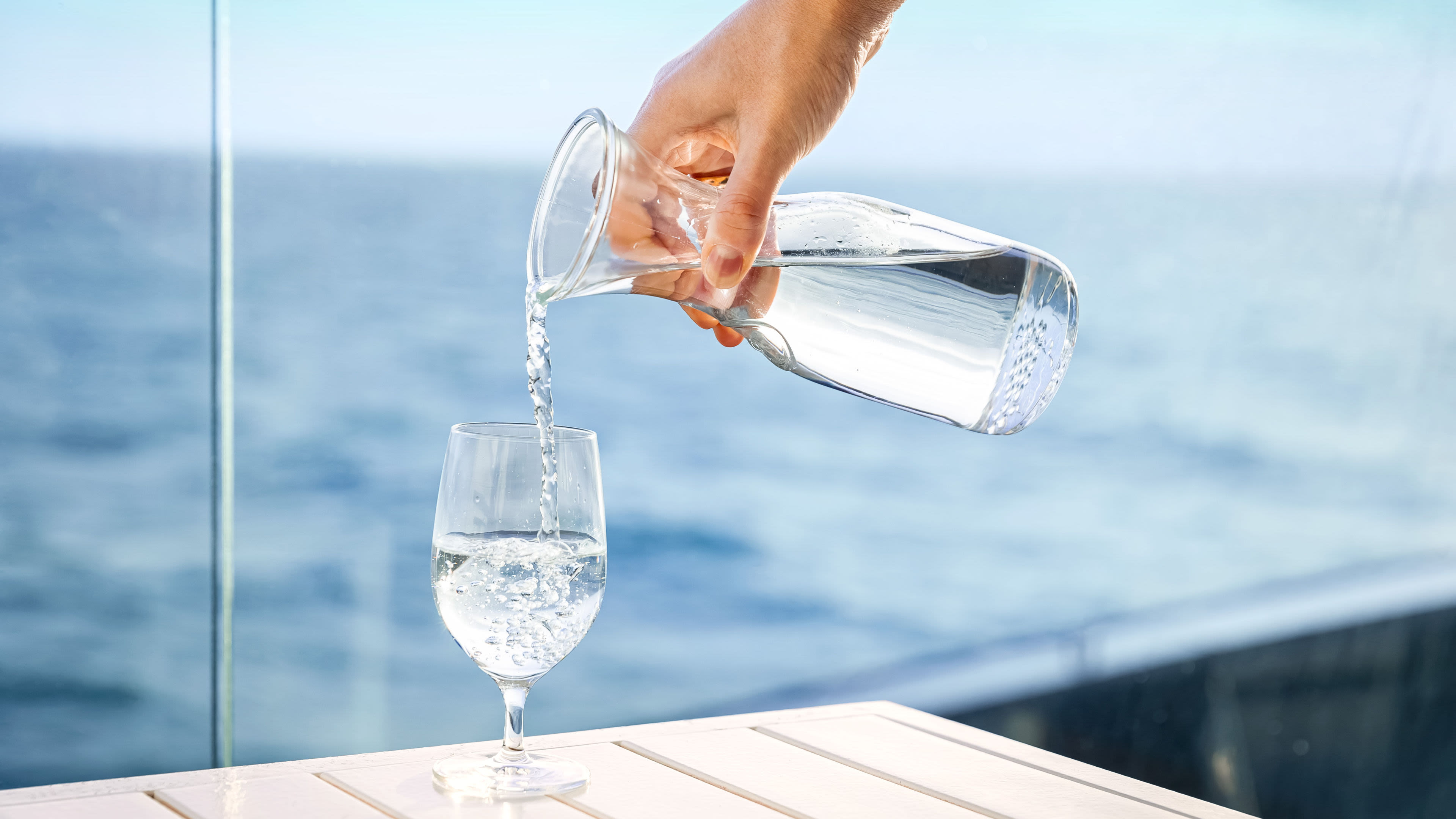 Hand gießt Wasser aus Karaffe ins Glas, Meereshintergrund.