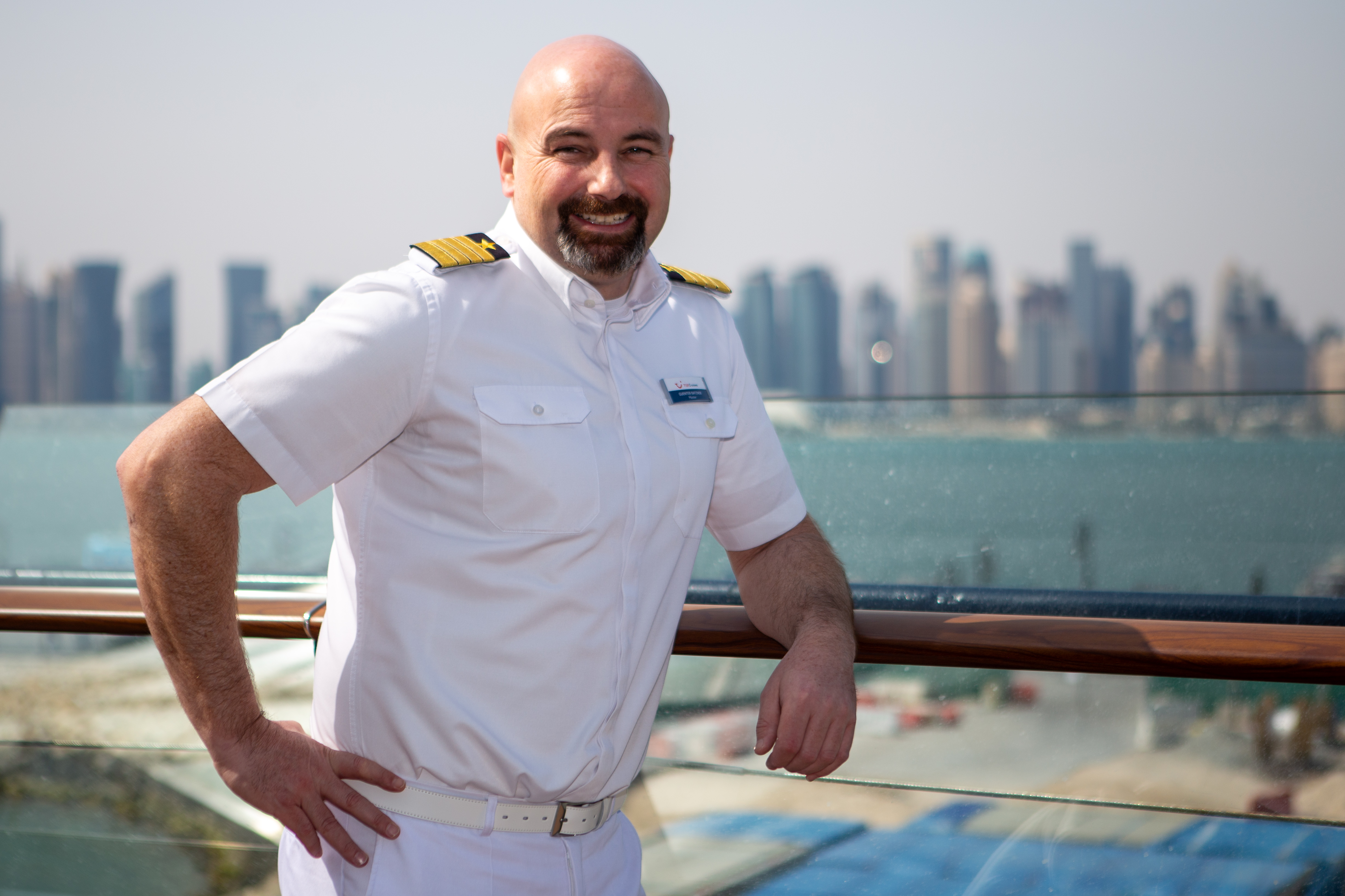 Mann in weißer Uniform auf Kreuzfahrtschiff, Skyline im Hintergrund.