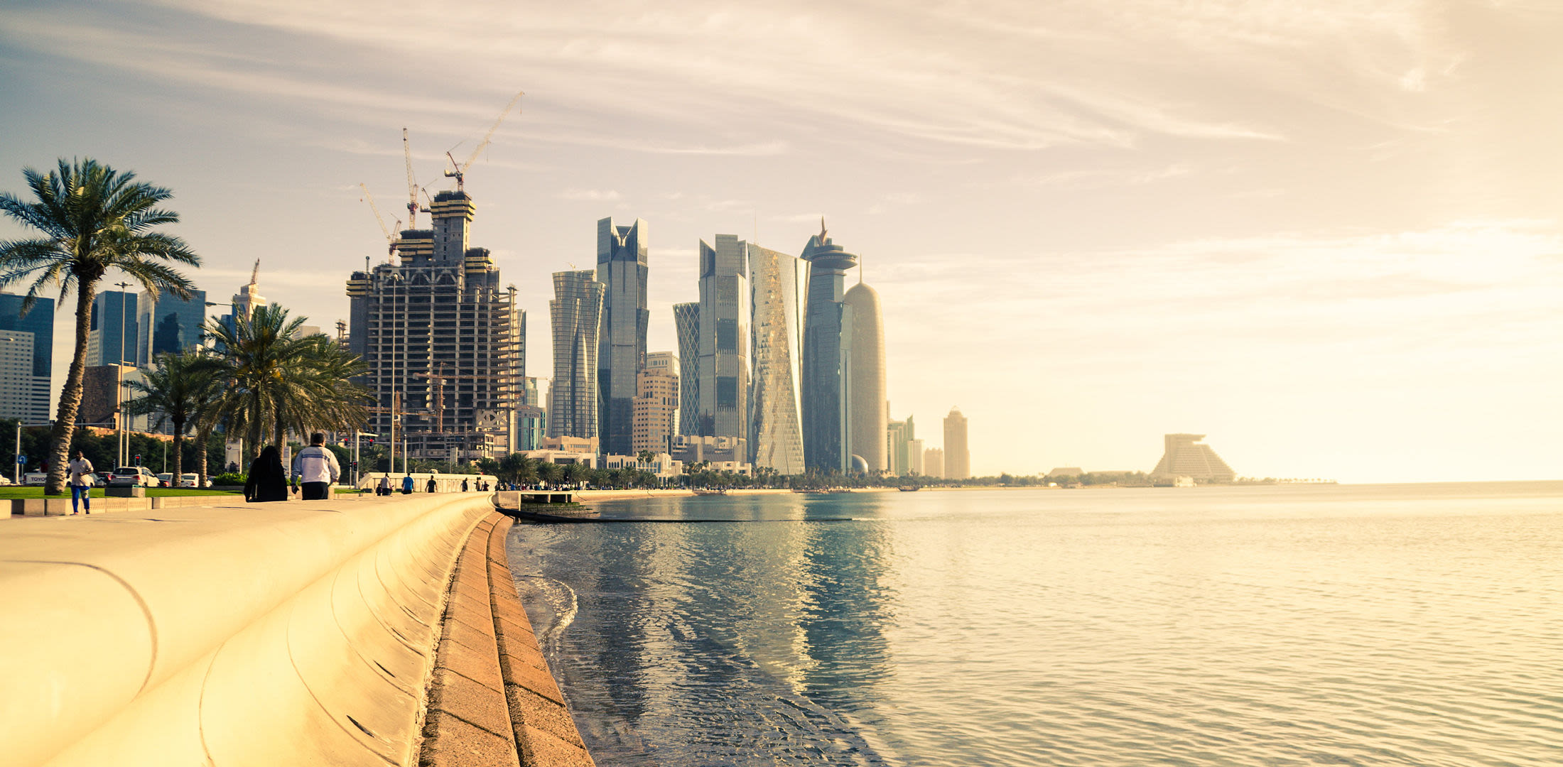 Doha Skyline bei Sonnenuntergang, Palmen, Wasser, Spaziergänger entlang der Küste.