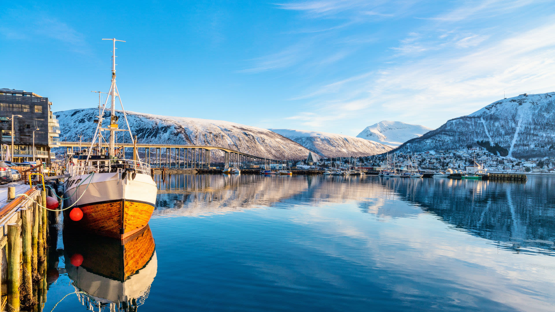 Holzboot im Hafen von Tromsø, Norwegen, mit schneebedeckten Bergen im Hintergrund.