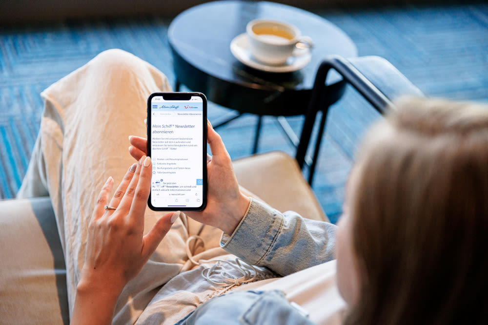 Frau liest Mein Schiff® Newsletter auf Smartphone, Kaffee im Hintergrund.