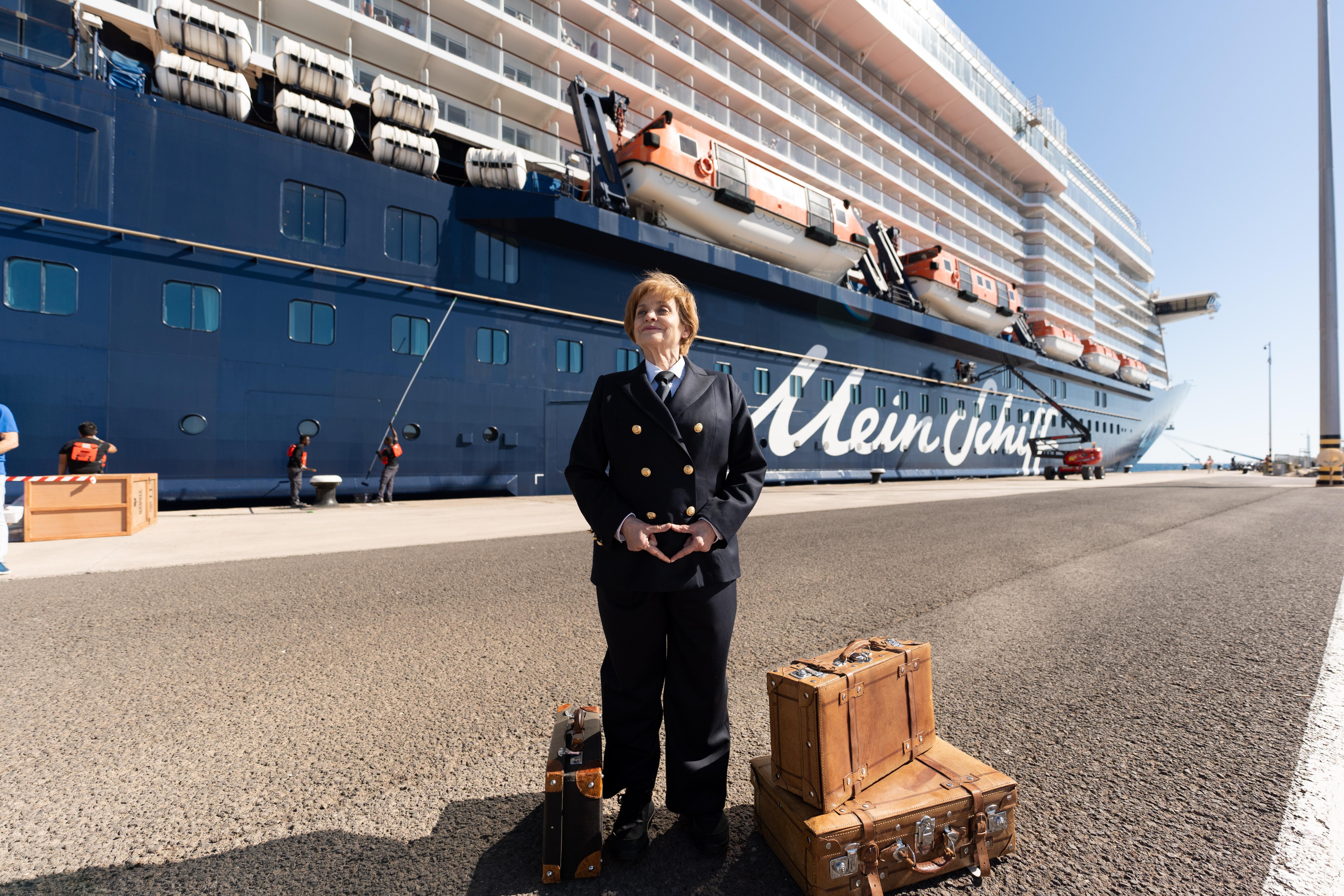 Frau in Uniform vor Kreuzfahrtschiff der Mein Schiff® Flotte, Koffer daneben.