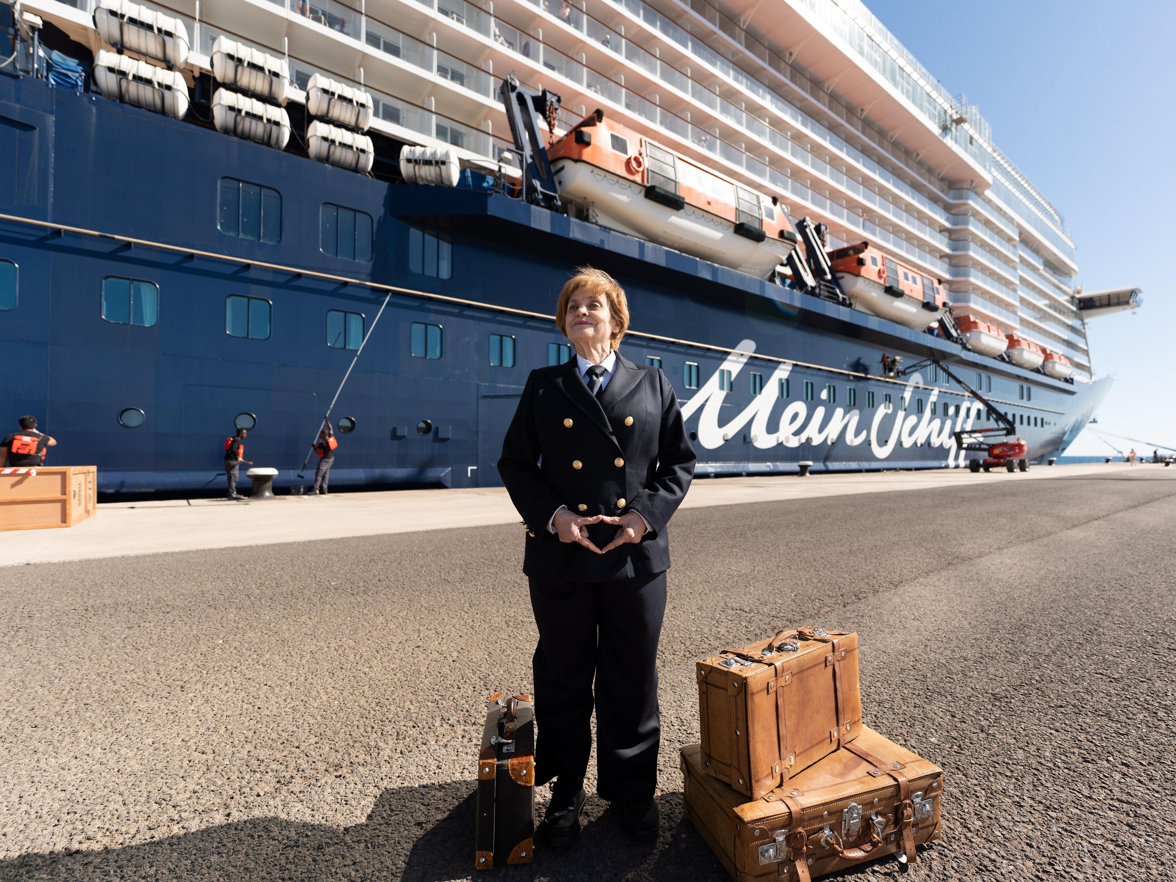 Miss Merkel - Mord auf hoher See Frau in Uniform vor Kreuzfahrtschiff der Mein Schiff® Flotte, Koffer daneben.