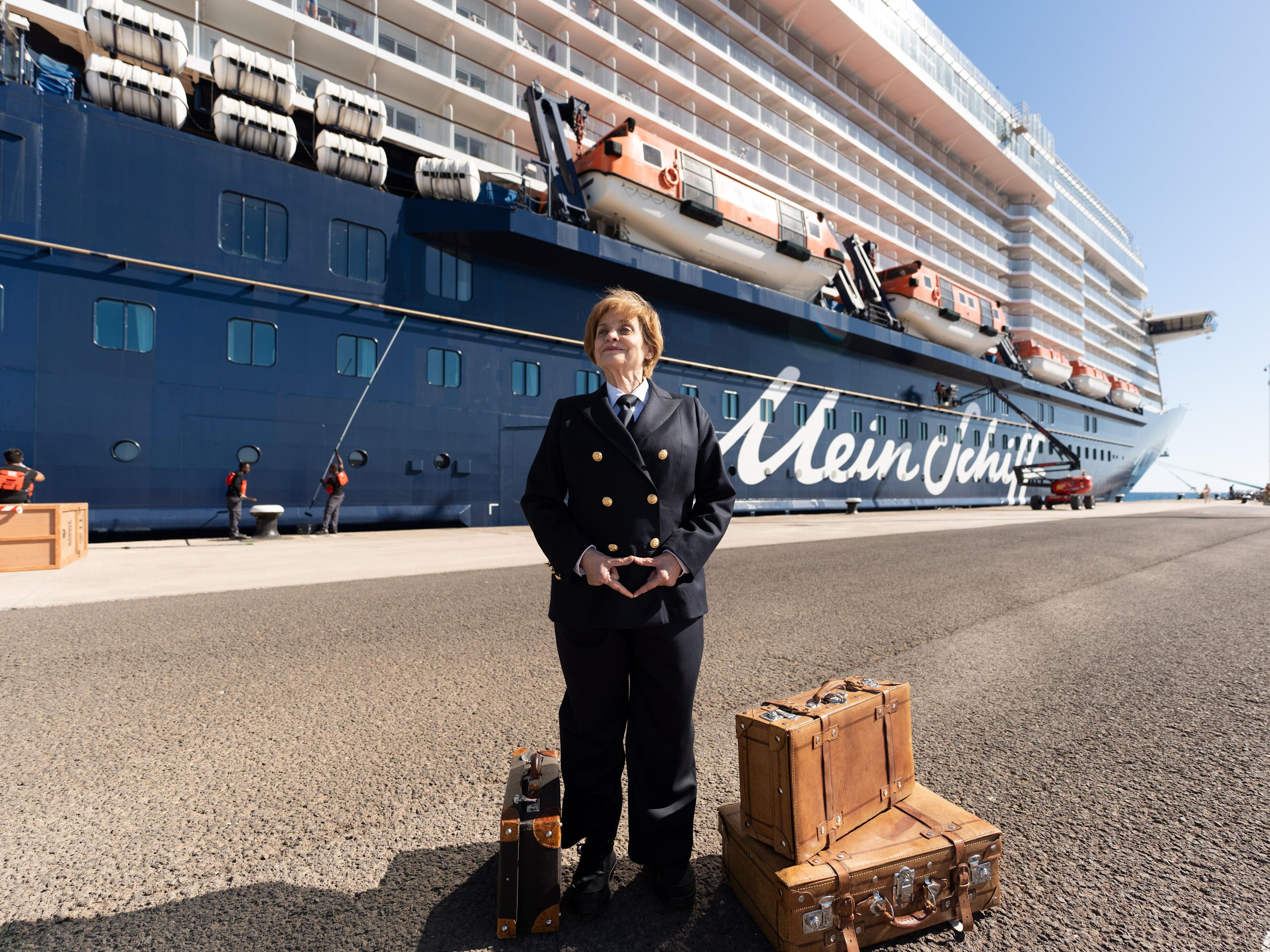 Frau in Uniform vor Kreuzfahrtschiff der Mein Schiff® Flotte, Koffer daneben. Frau in Uniform vor Kreuzfahrtschiff der Mein Schiff® Flotte, Koffer daneben.