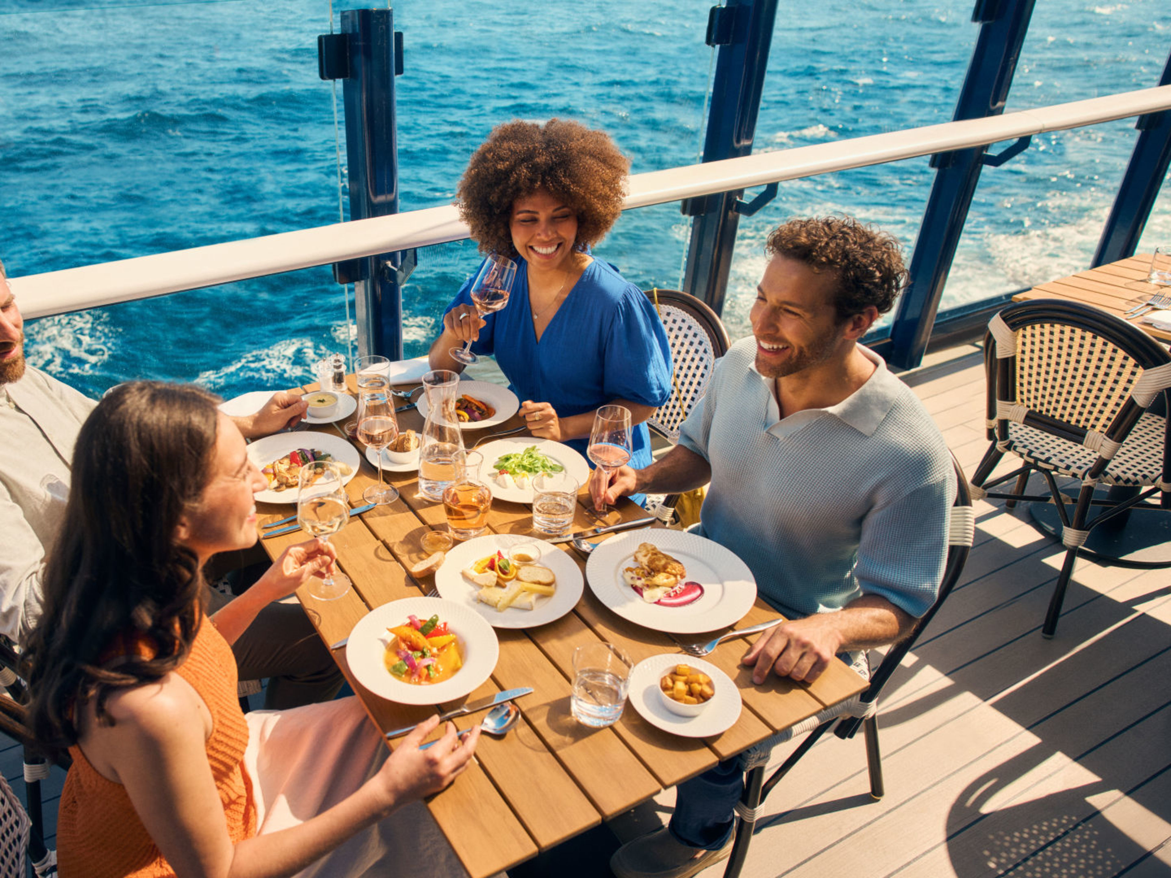 Freundesgruppe genießt ihr Essen im Außenbereich des Sur Mer Bistrots Vier Personen essen auf einem Kreuzfahrtschiff der Mein Schiff® Flotte mit Meerblick.