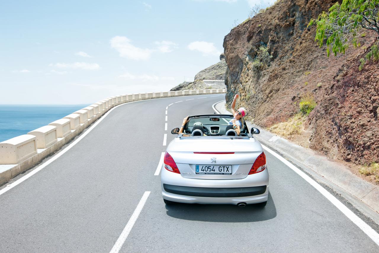 Mietwagen Silbernes Cabrio auf Küstenstraße, zwei Personen, sonniger Tag, Meerblick.