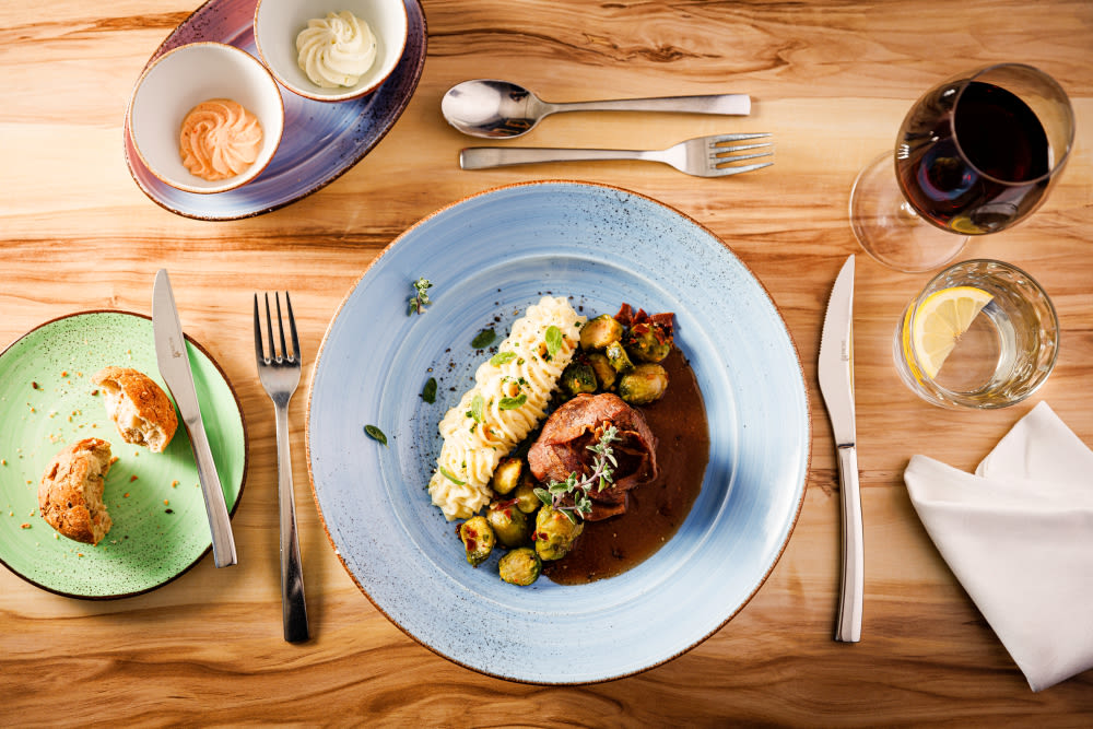 Gedeckter Tisch mit Fleischgericht, Kartoffelpüree, Rosenkohl, Rotwein und Wasser.