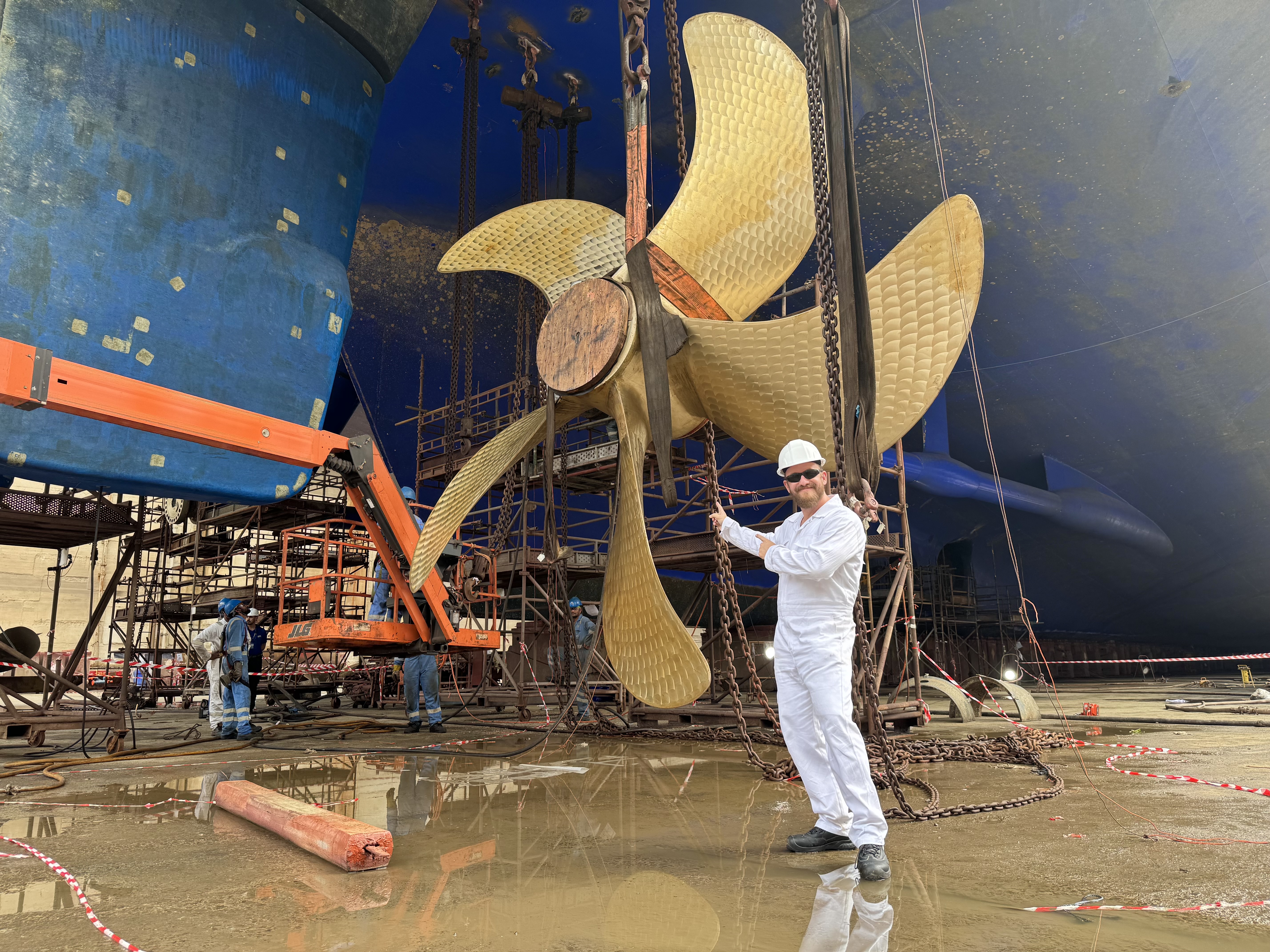 Mann in weißem Overall vor großem Schiffspropeller in Werft.